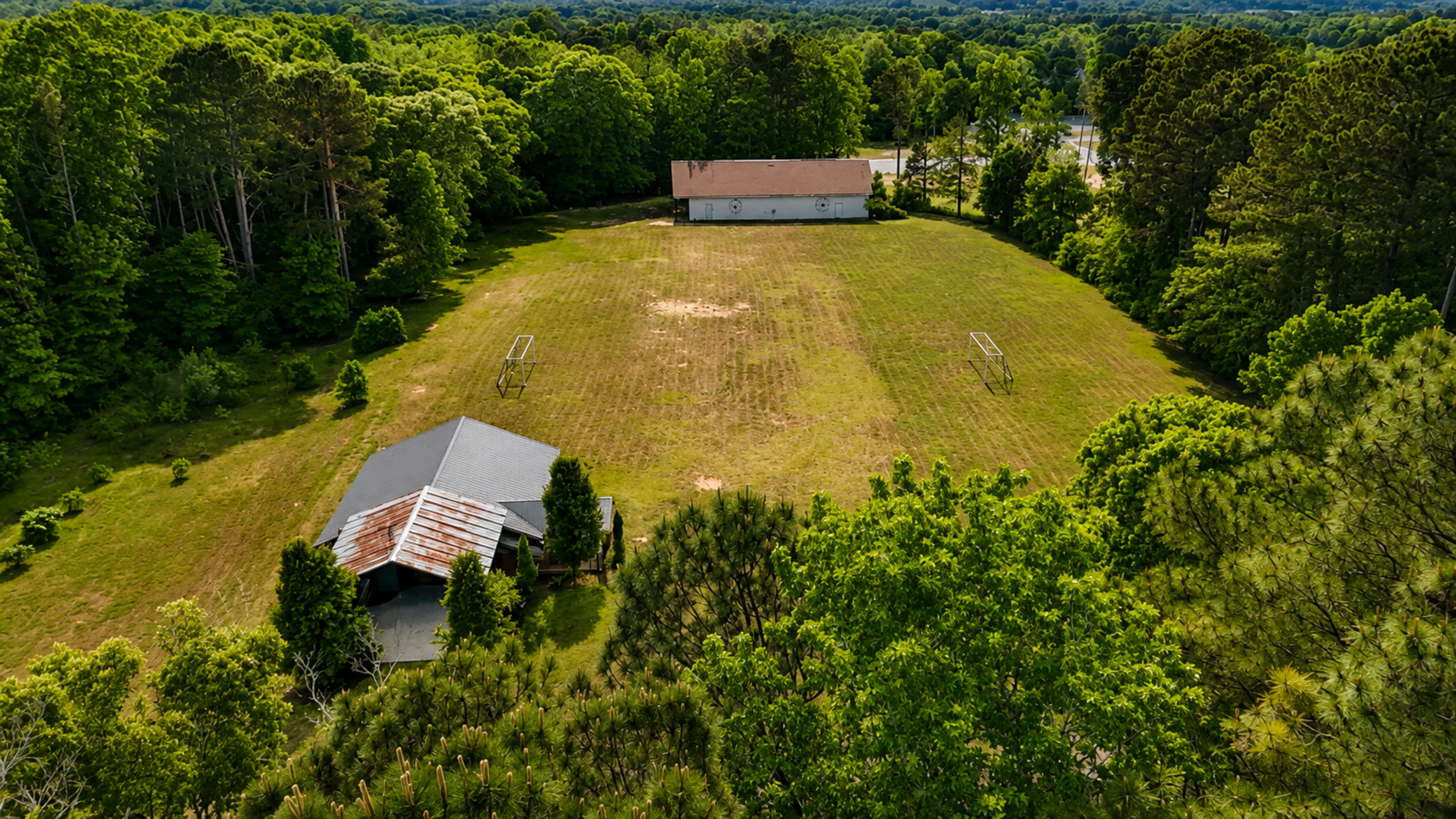 The image shows an expansive grassy area surrounded by trees, with two buildings, one of which has a red-tinted roof, positioned on the landscape.