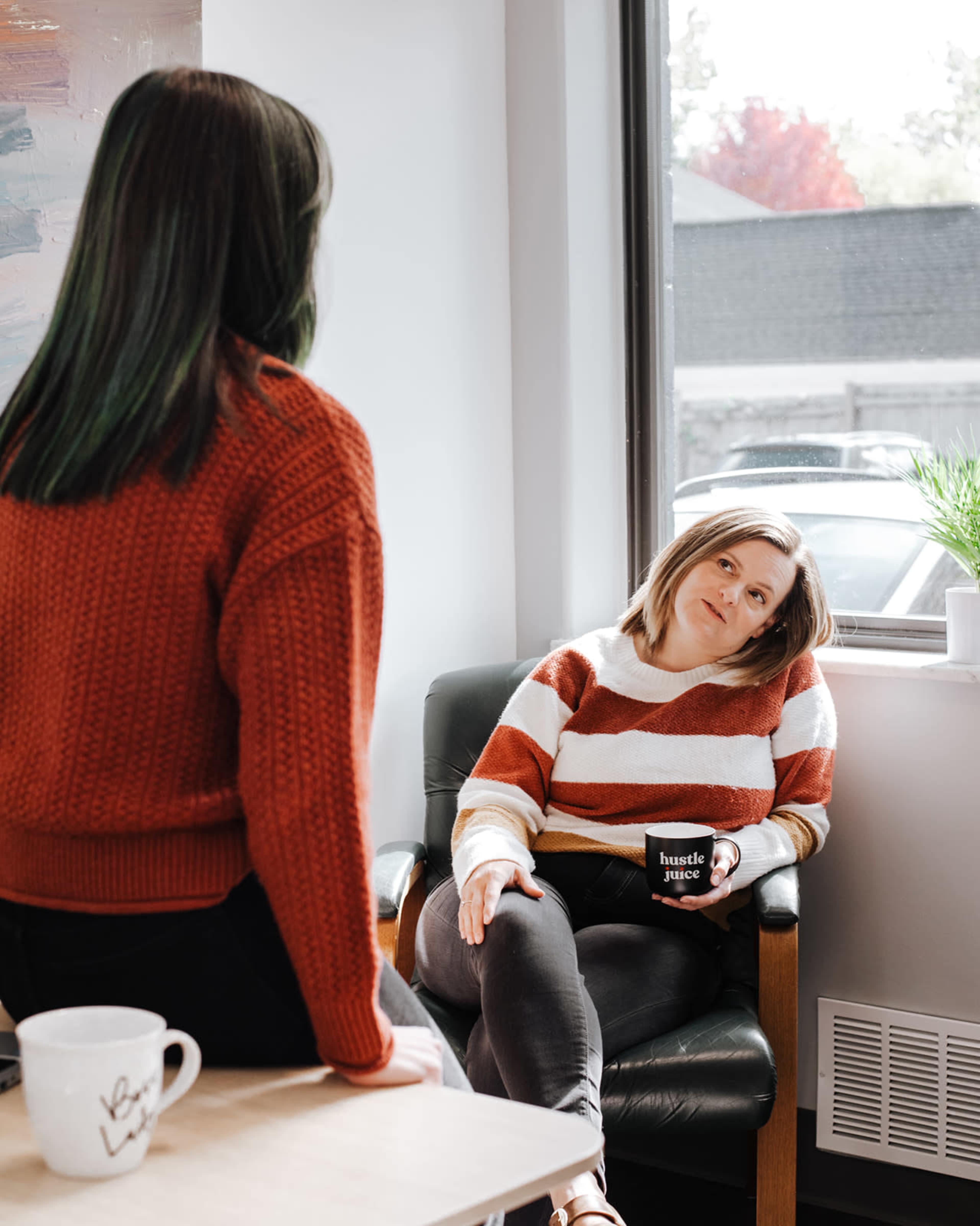 Two women sit in a brightly lit room, one seated in an armchair with a mug, while the other stands nearby, both wearing sweaters.