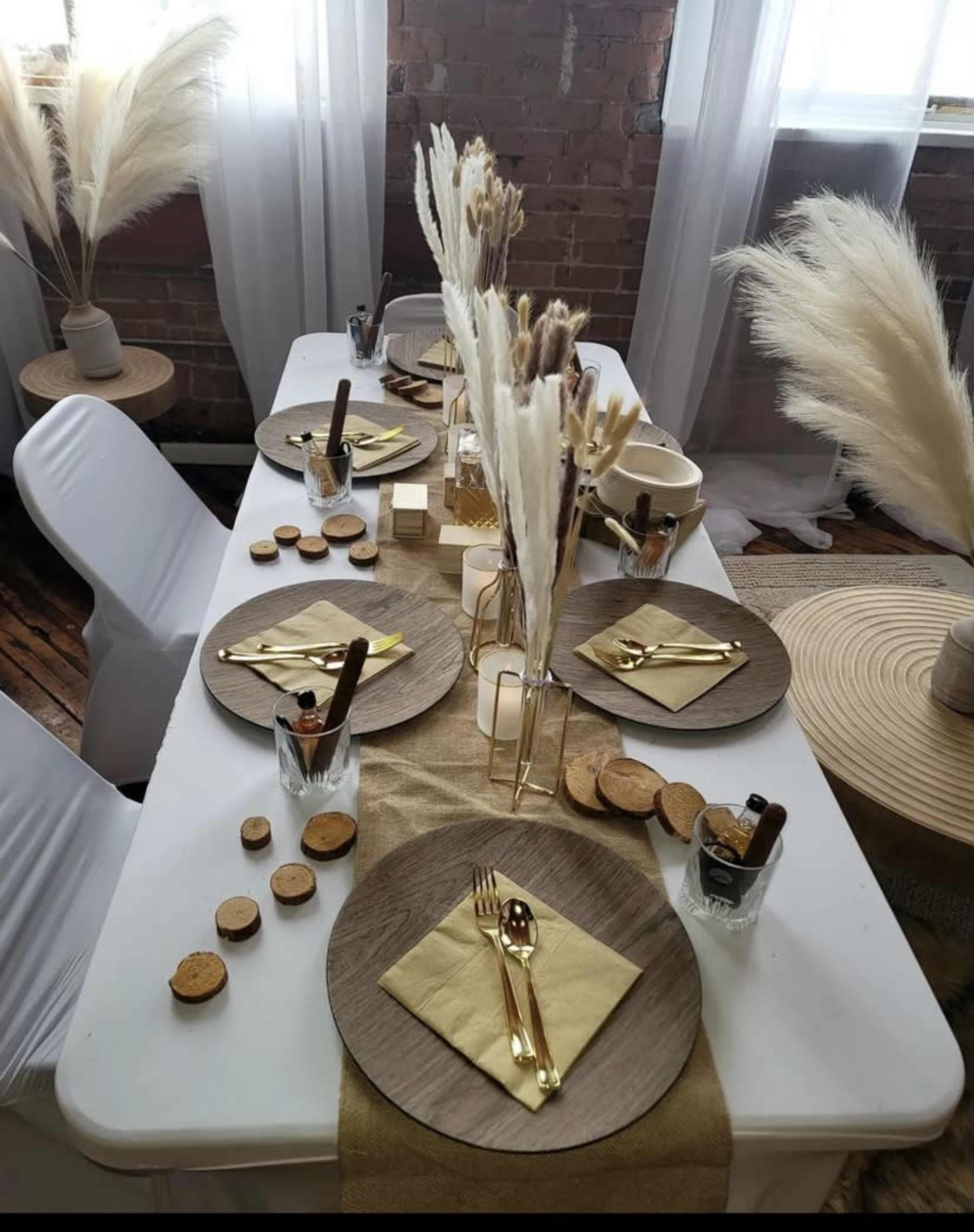 A dining table is elegantly set with neutral-colored plates, cutlery, and decorative dried florals, surrounded by simple chairs and wooden coasters.