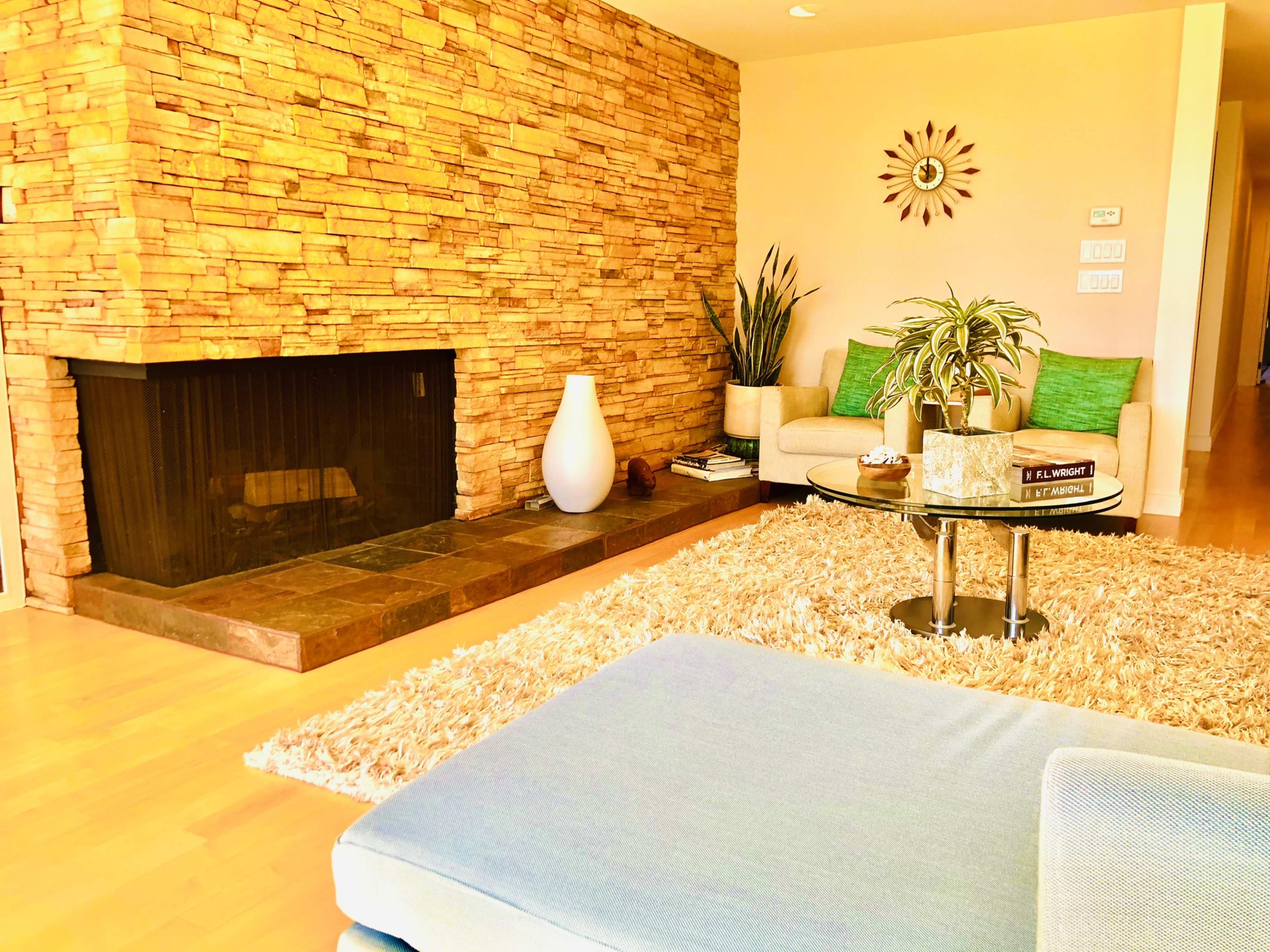 A modern living room with a stone fireplace, a round glass coffee table, a large area rug, and a comfortable seating arrangement against a light-colored wall.