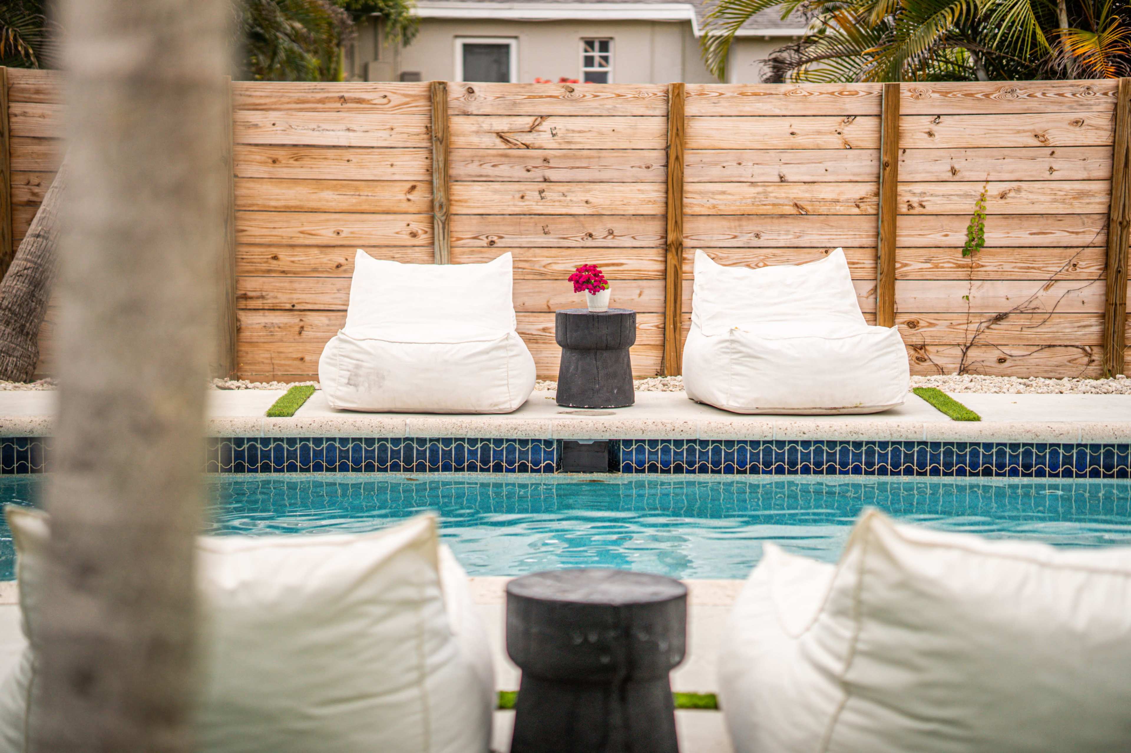 The image shows a tranquil pool area featuring two white bean bag chairs positioned by the edge of the pool, with a small table holding a flower between them, set against a wooden fence.