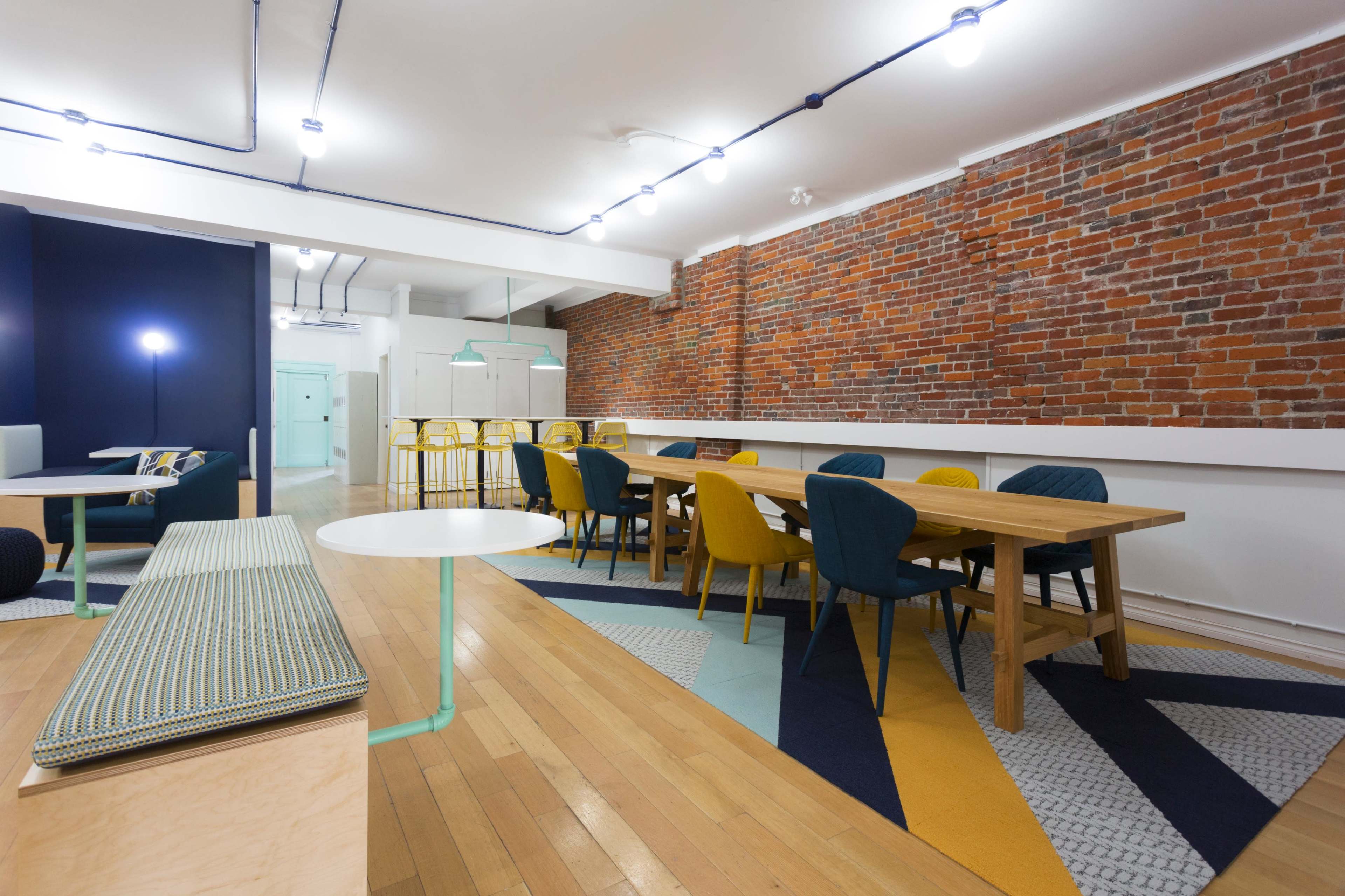 A modern communal space with a wooden dining table surrounded by colorful chairs, a patterned rug, and exposed brick walls.