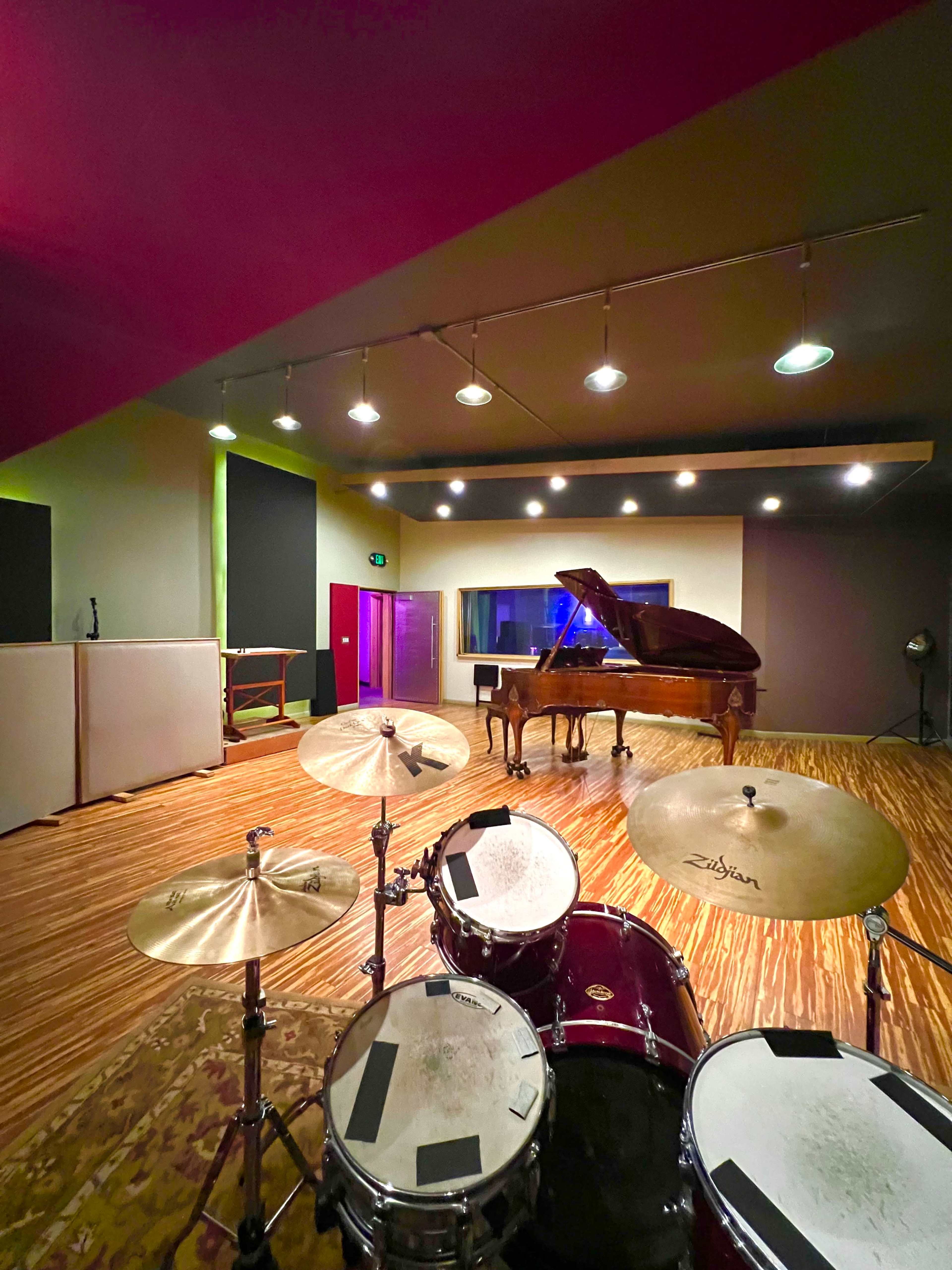 The image shows a music recording studio with a drum set in the foreground and a grand piano in the background.