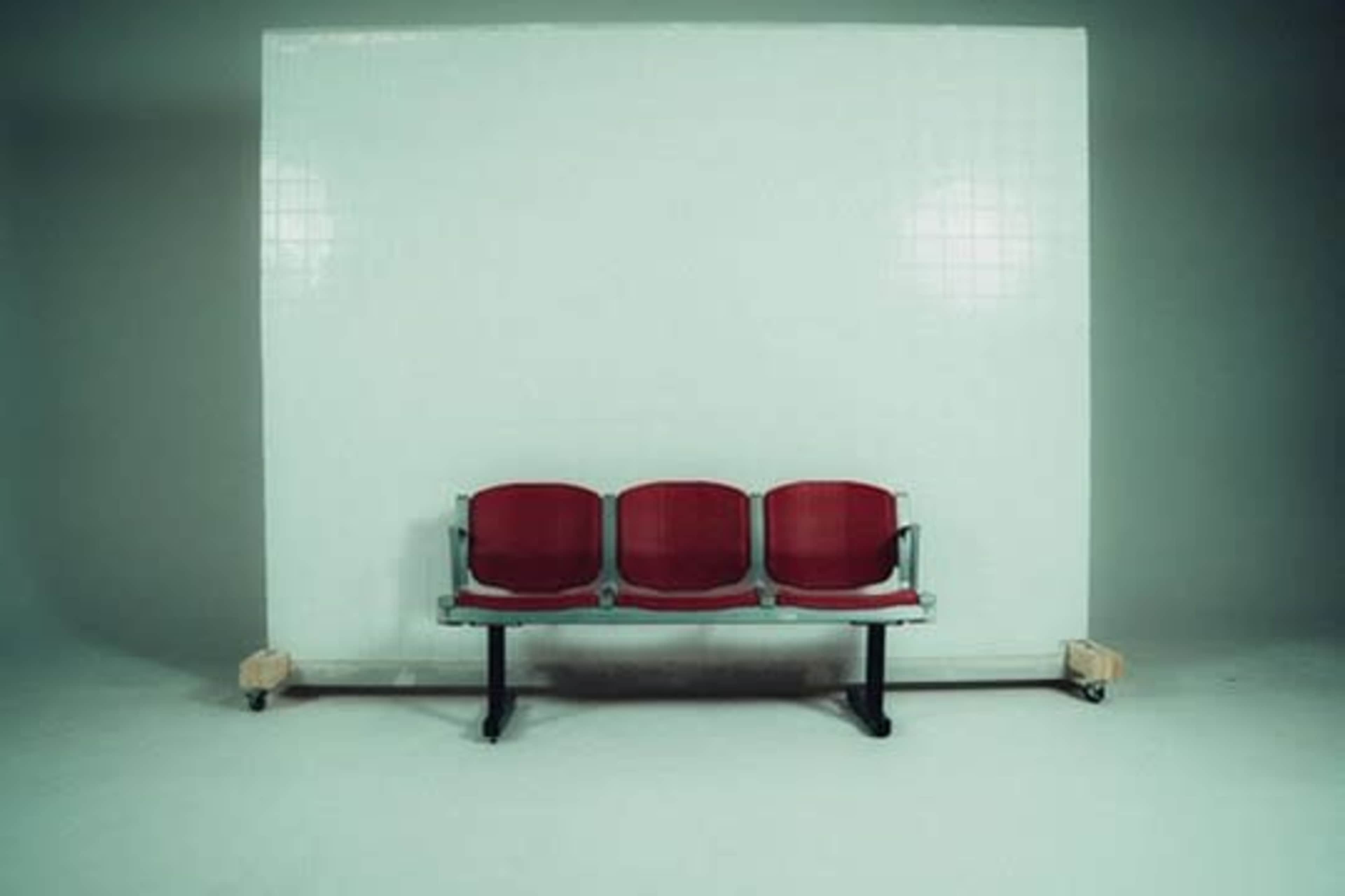The image shows a row of three red waiting room chairs against a plain white wall on a minimalist surface.