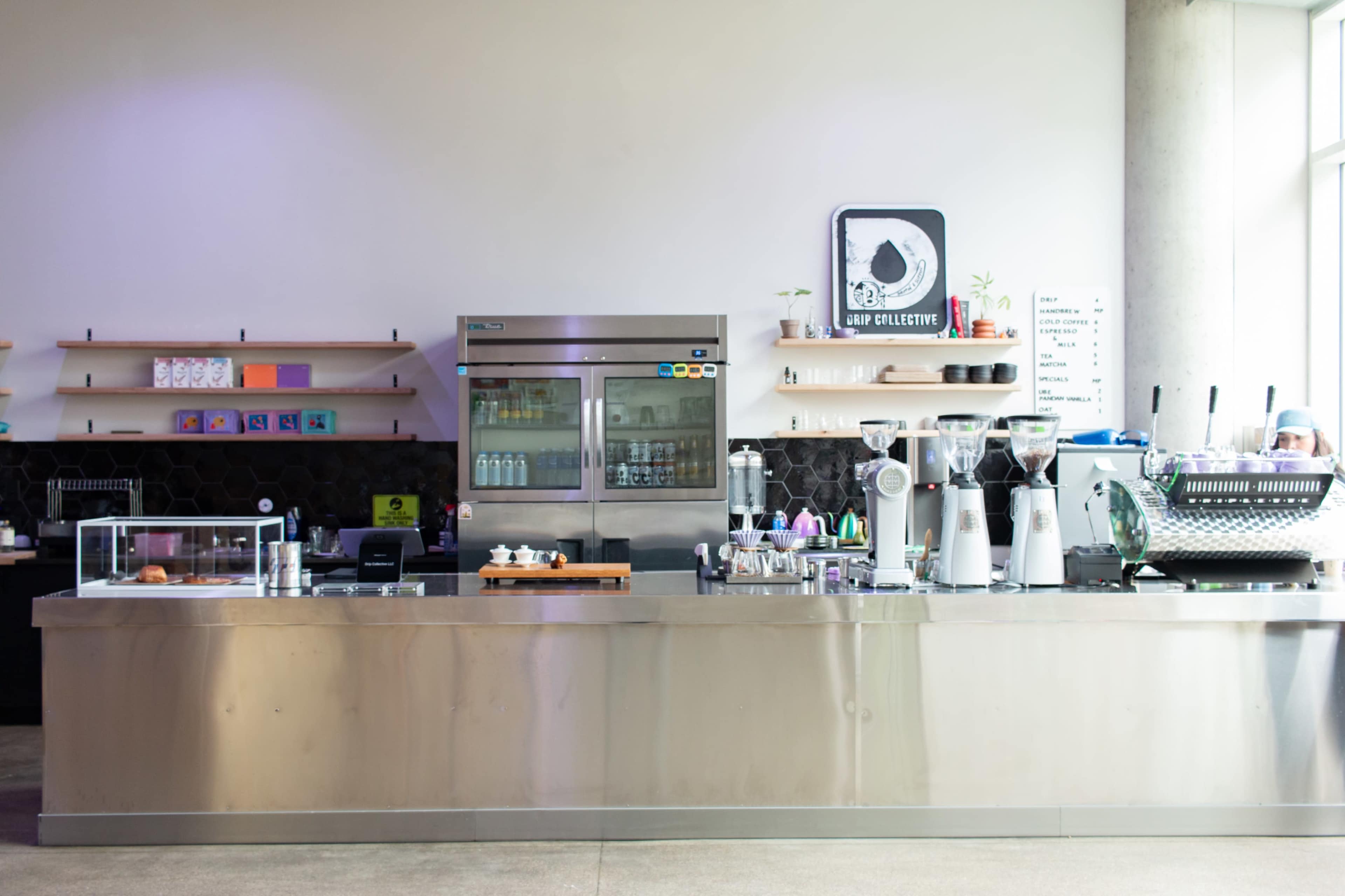 The image shows a modern café counter with a stainless steel surface, glass display case, coffee machines, and shelf-mounted products.