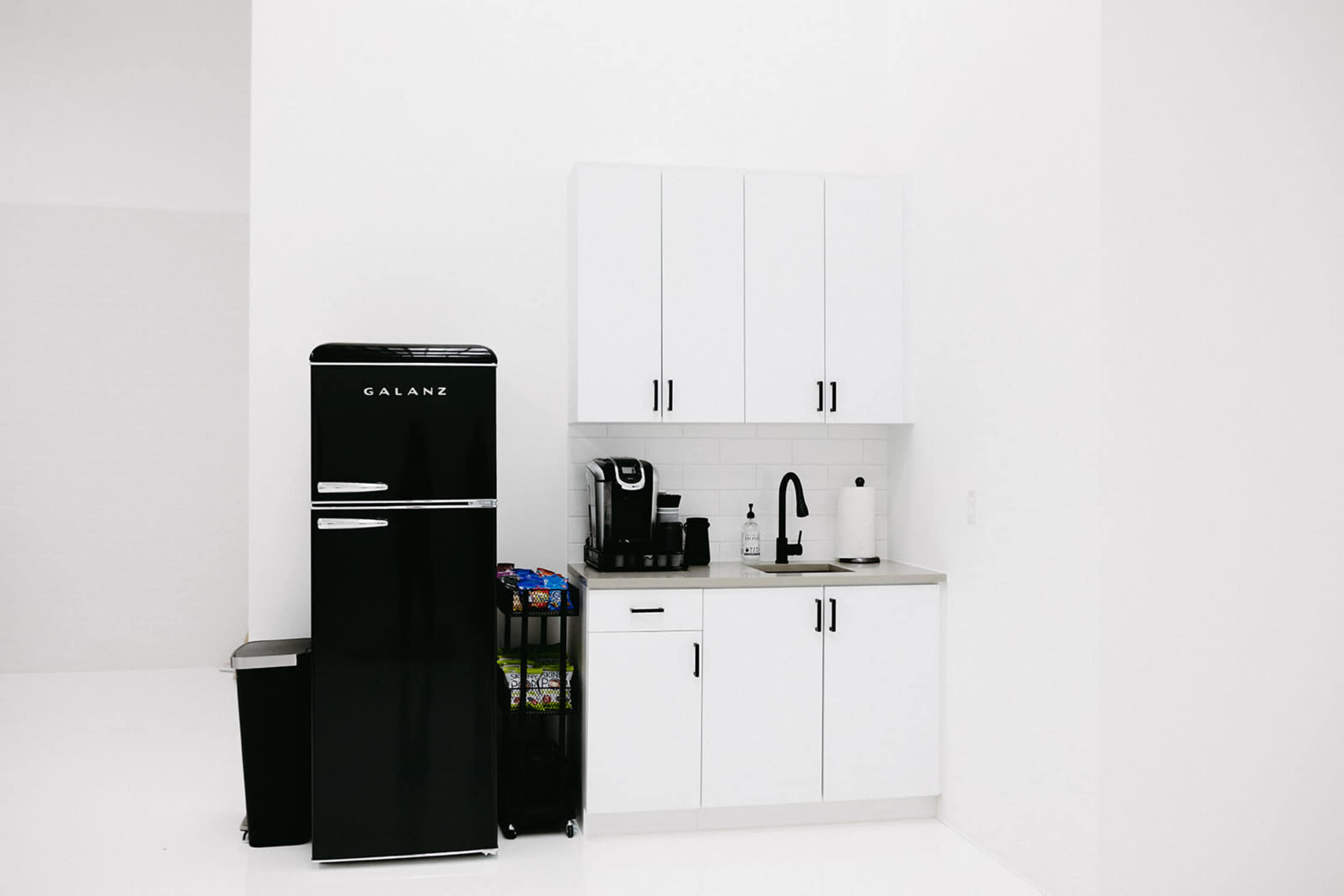 A minimalistic kitchen setup with a black refrigerator, a coffee maker, and white cabinets against a bright backdrop.