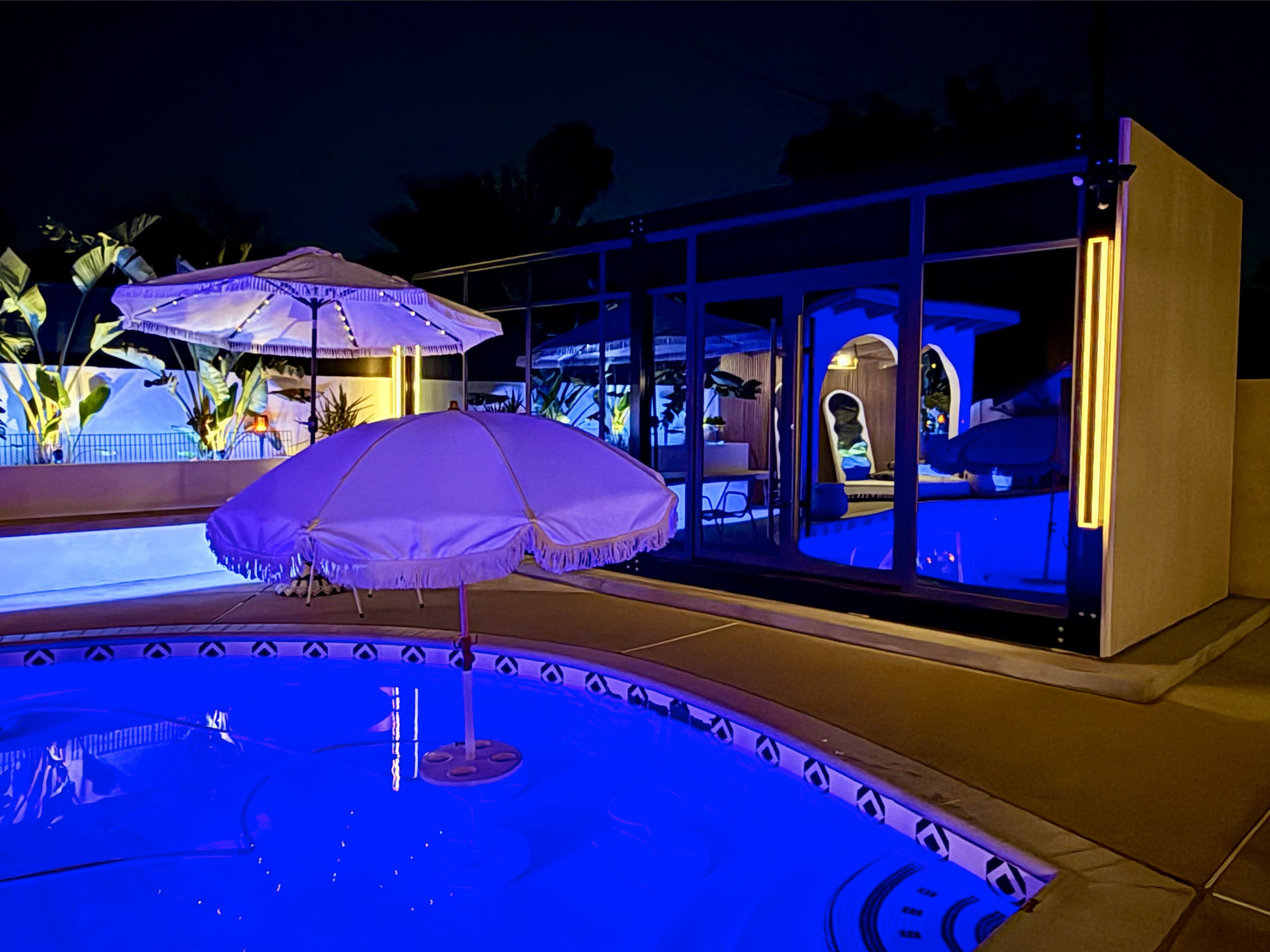 The image shows a swimming pool area at night, featuring illuminated water, an umbrella, and a modern structure in the background.