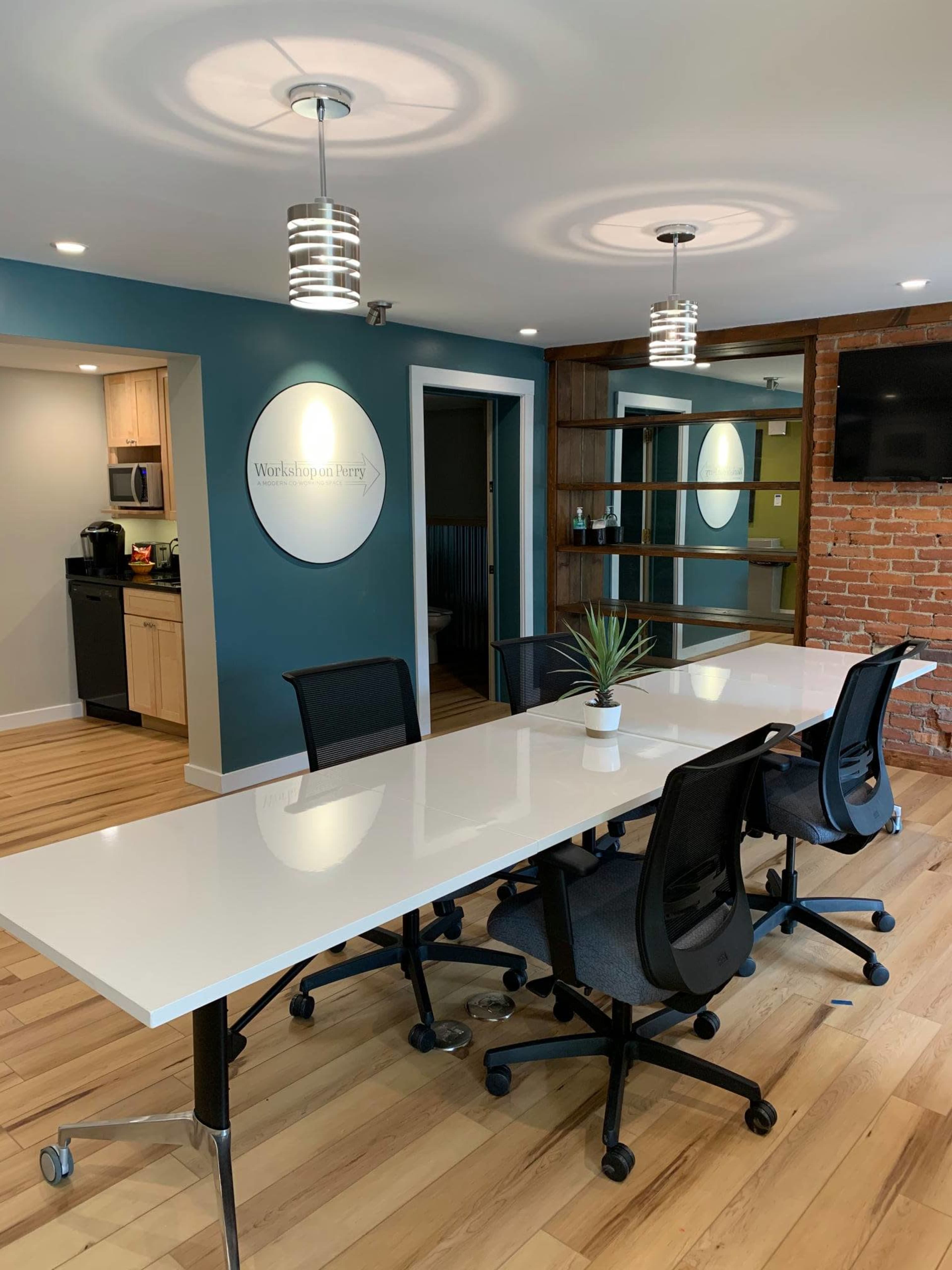 A modern conference room features a large white table surrounded by black chairs, with a brick wall and shelving in the background.