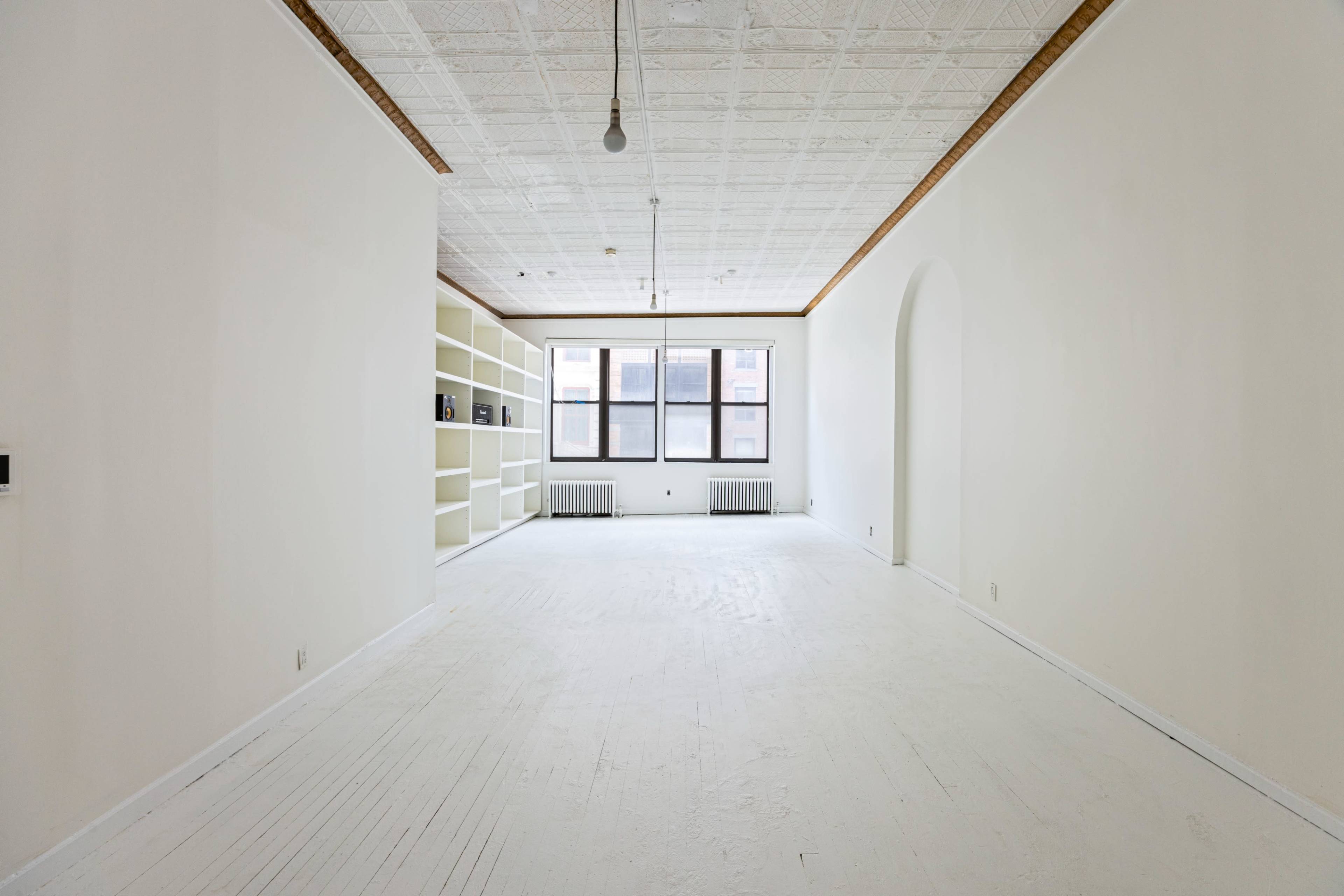 The image shows a spacious, empty room with white walls and a polished floor, featuring large windows and a built-in bookshelf along one wall.