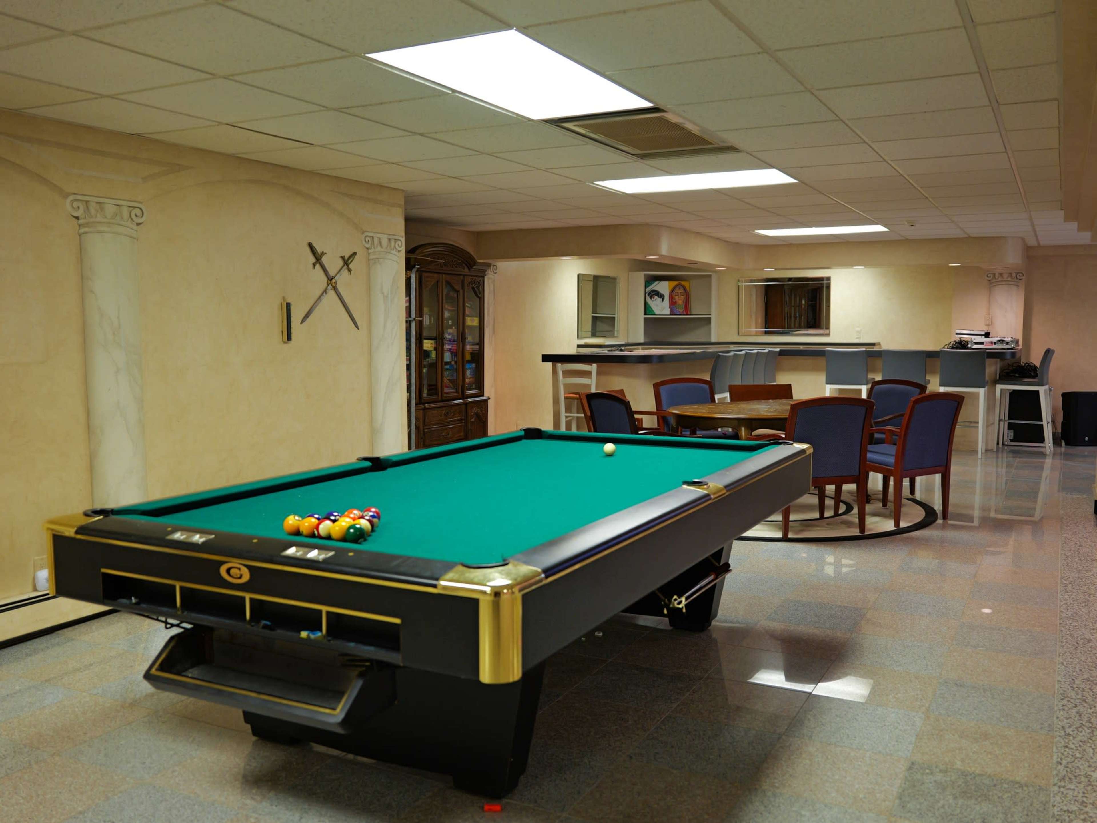 The image shows a game room featuring a green felt pool table, a dining area with chairs, and a counter in the background.