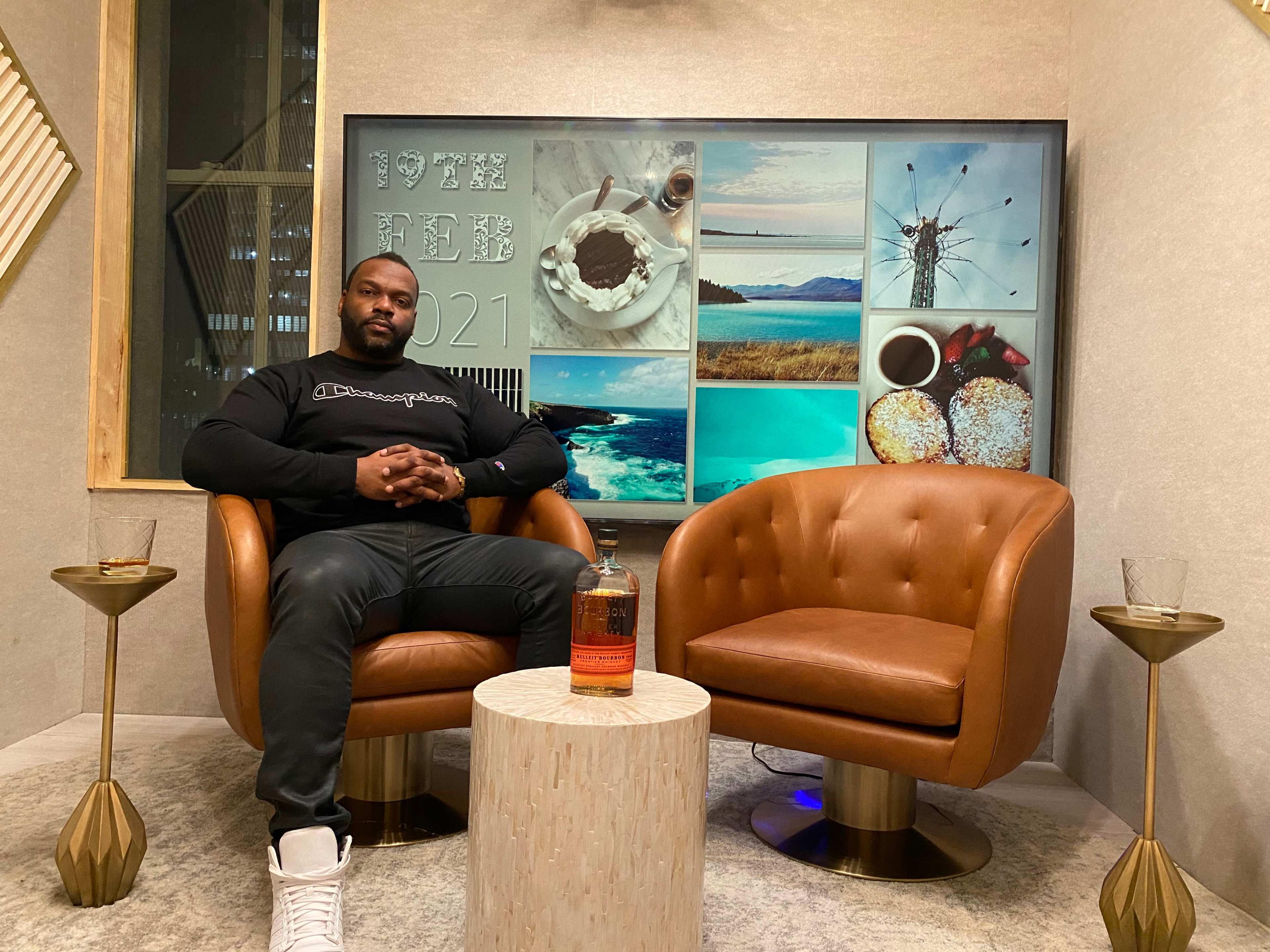 A man is seated in a cozy setting with two brown chairs and a decorative wall featuring various images, alongside a bottle of whiskey on a small table.