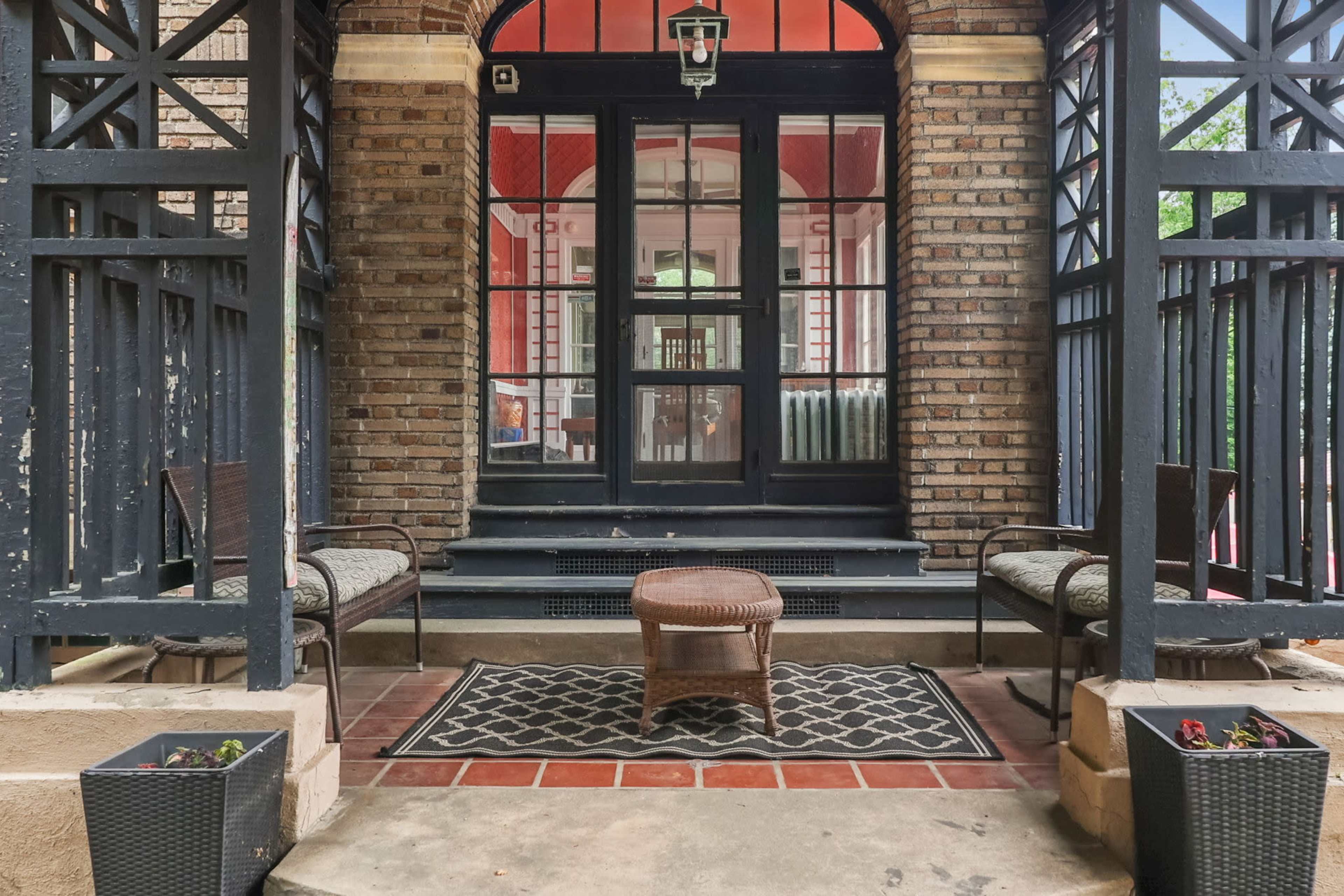A porch area with two chairs, a small table, and potted plants, leading to a glass door framed by brick walls.