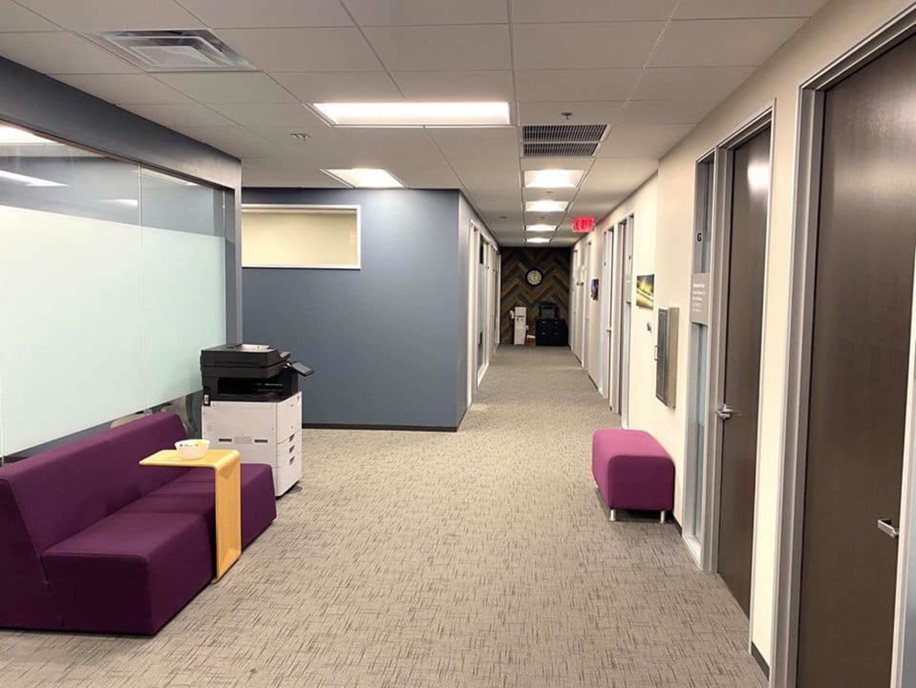 A clean office hallway with purple seating, light gray carpet, and doors leading to individual offices.