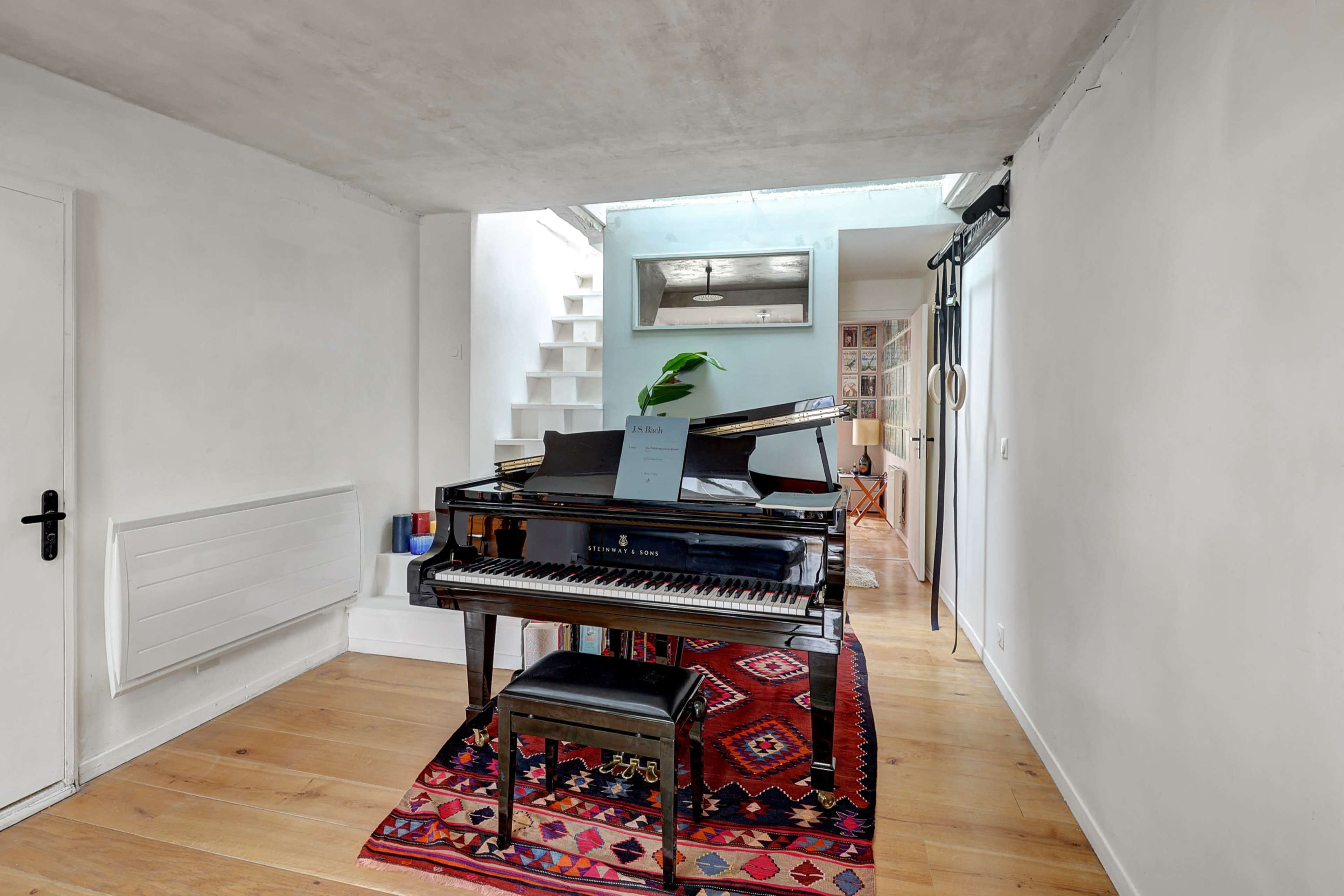 A black piano sits on a colorful rug in a bright room with white walls and a staircase in the background.