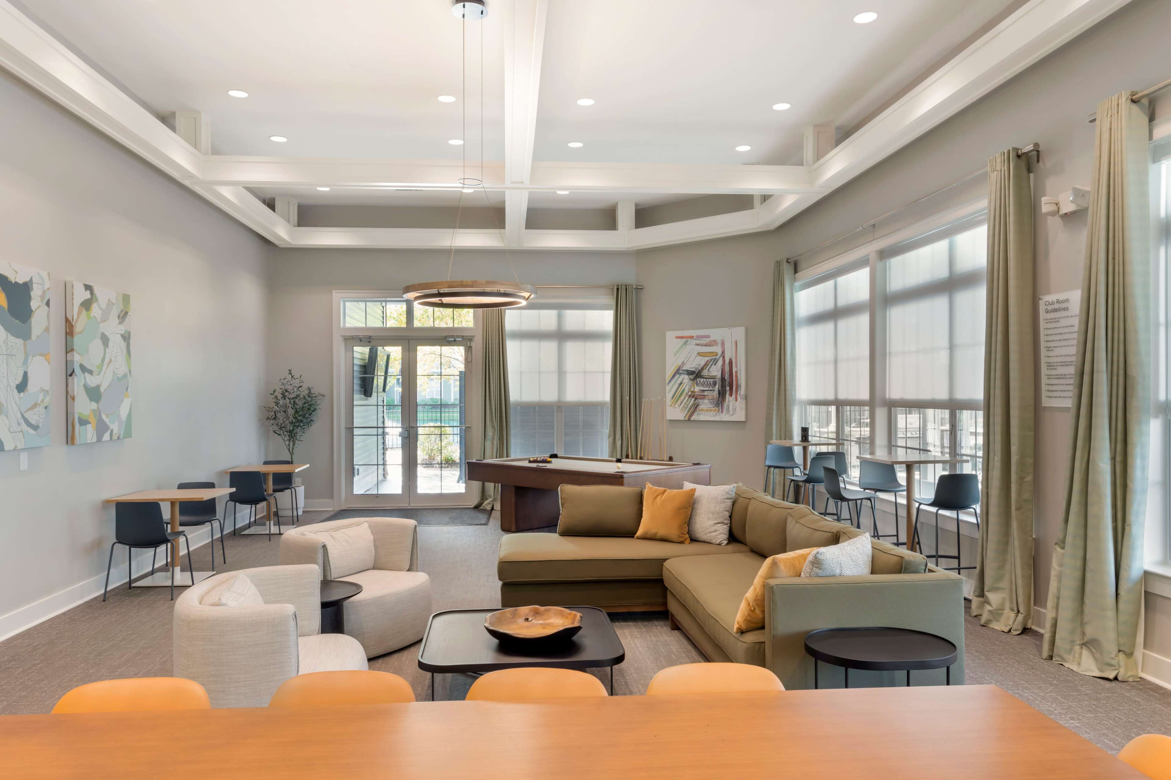 The image shows a spacious, modern lounge area with a sectional sofa, a pool table, seating for several people, and large windows letting in natural light.