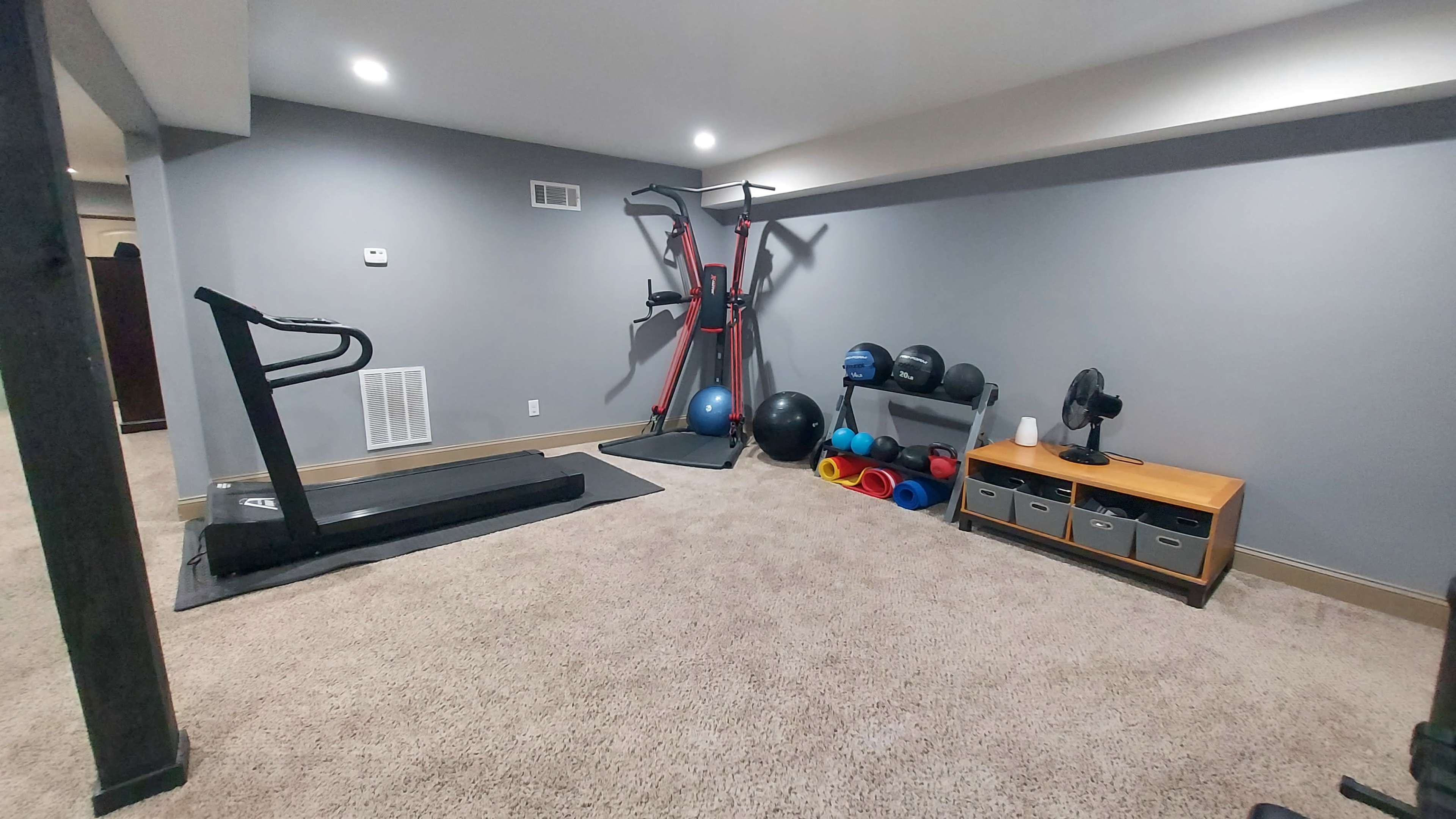 A home gym with a treadmill, a multi-gym machine, various exercise equipment, and a wooden storage unit against the wall.