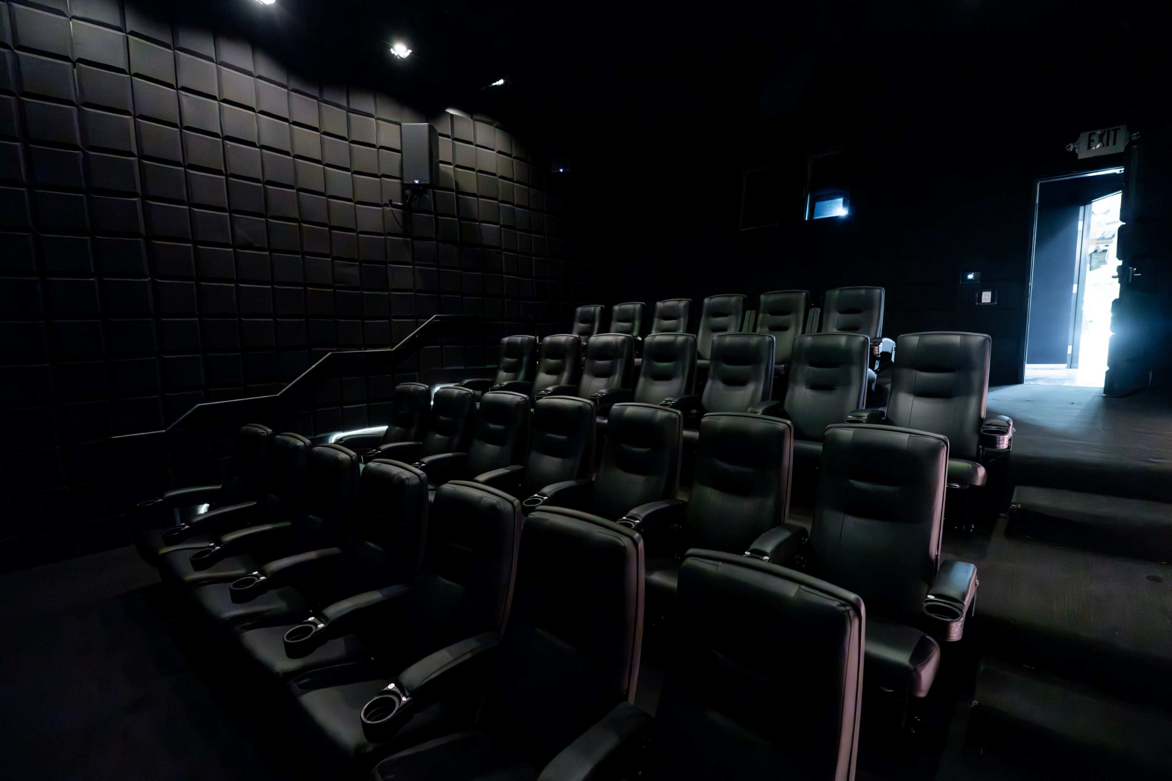 The image shows a dark theater with a row of black leather seating and an exit door in the background.