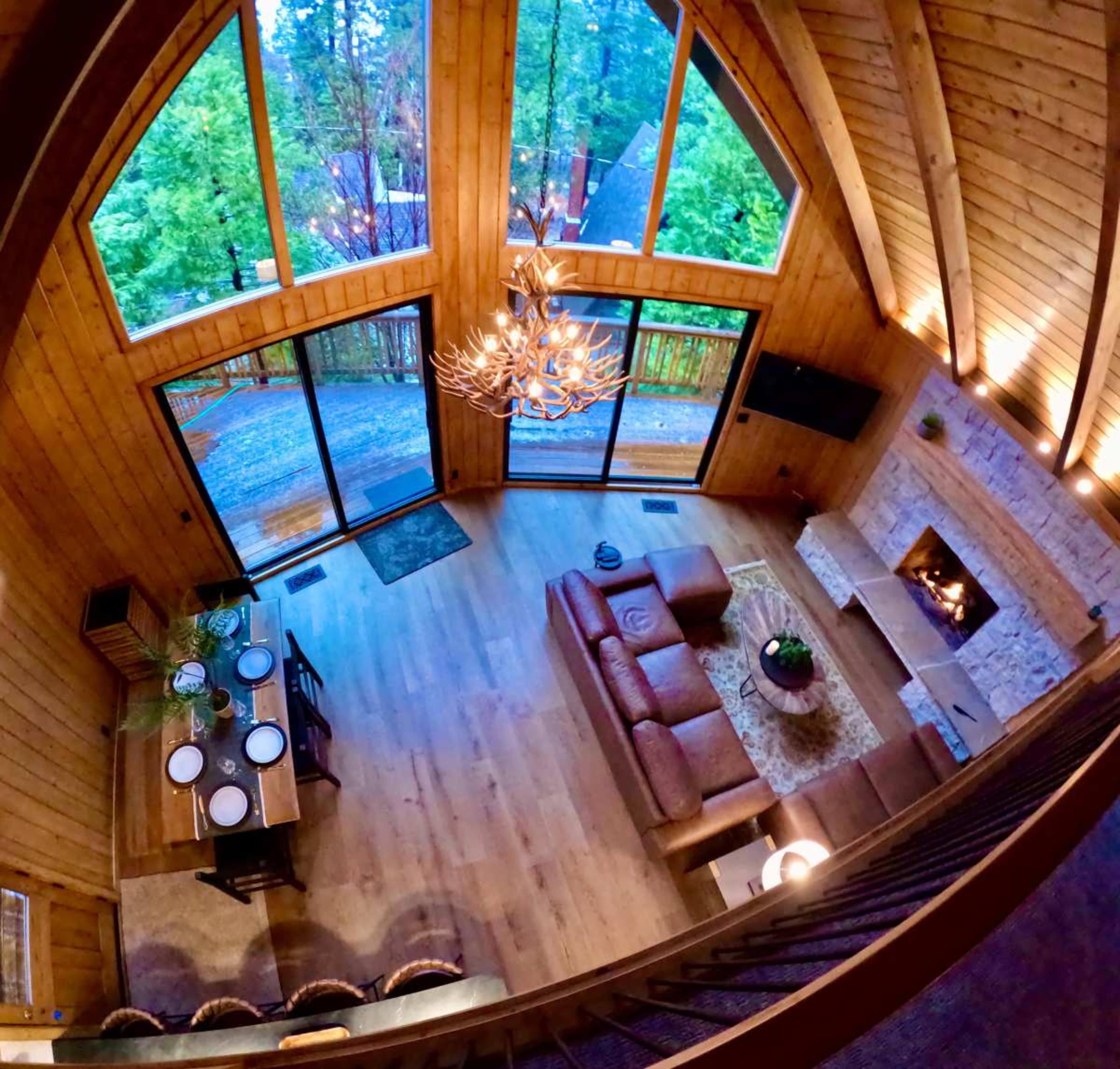 The image shows a spacious wooden cabin interior with large windows, a dining table, a leather couch, and a stone fireplace under a high ceiling.