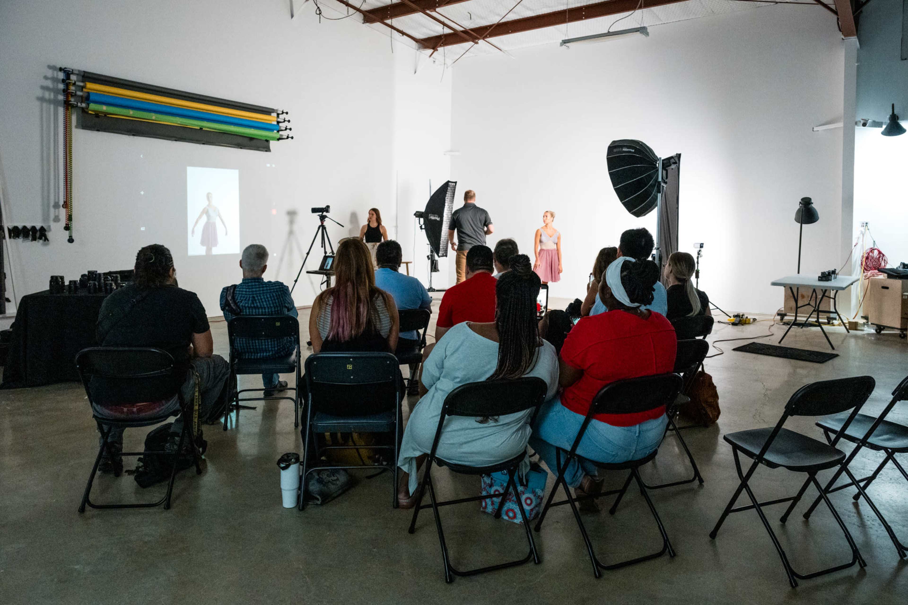 A group of individuals sits in chairs facing a stage with a model and photographers, while equipment is set up for a photo shoot in a studio.