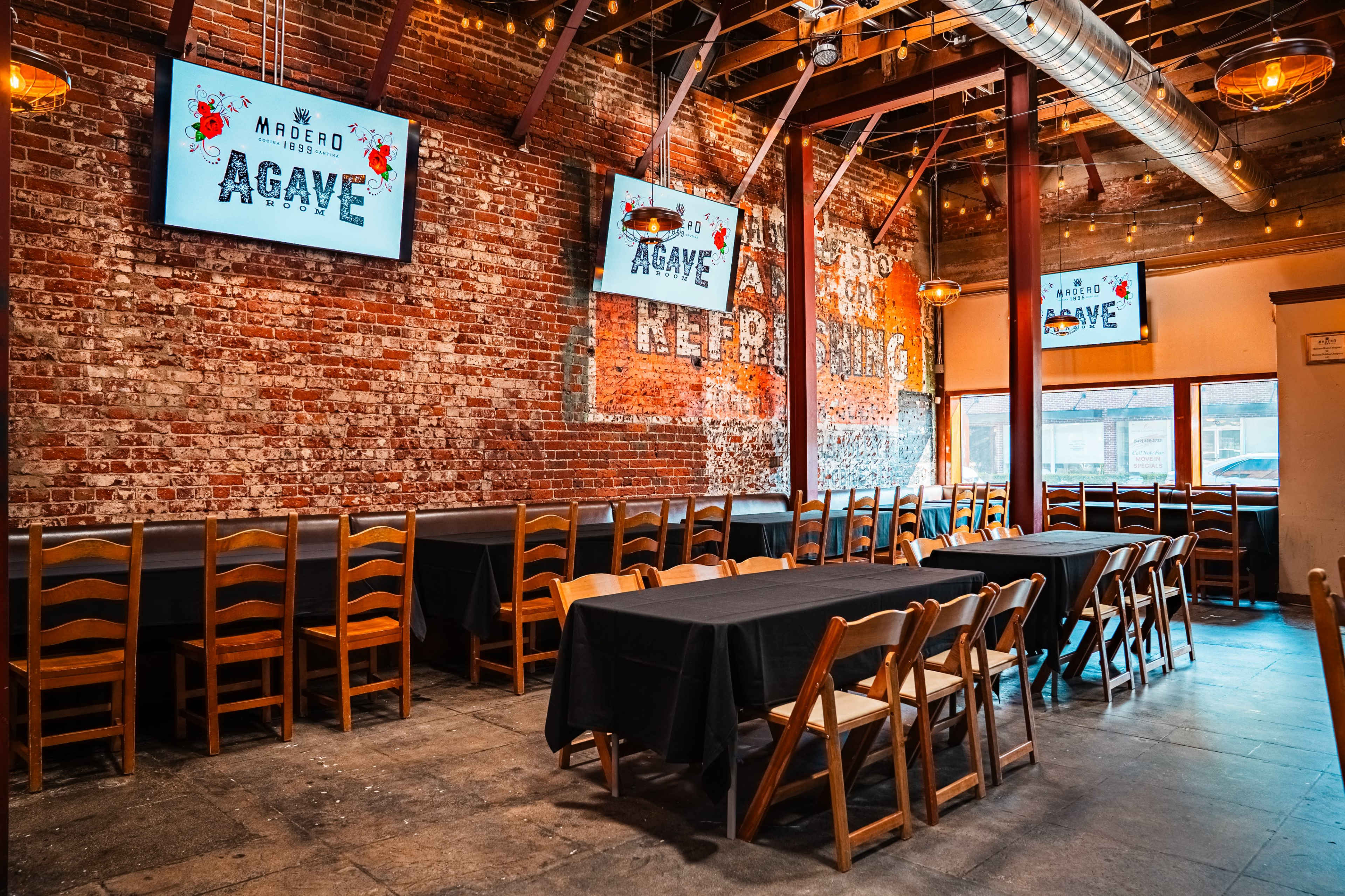 The image shows a spacious restaurant interior with exposed brick walls, wooden beams, and several long tables set up under illuminated signage for "Agave."