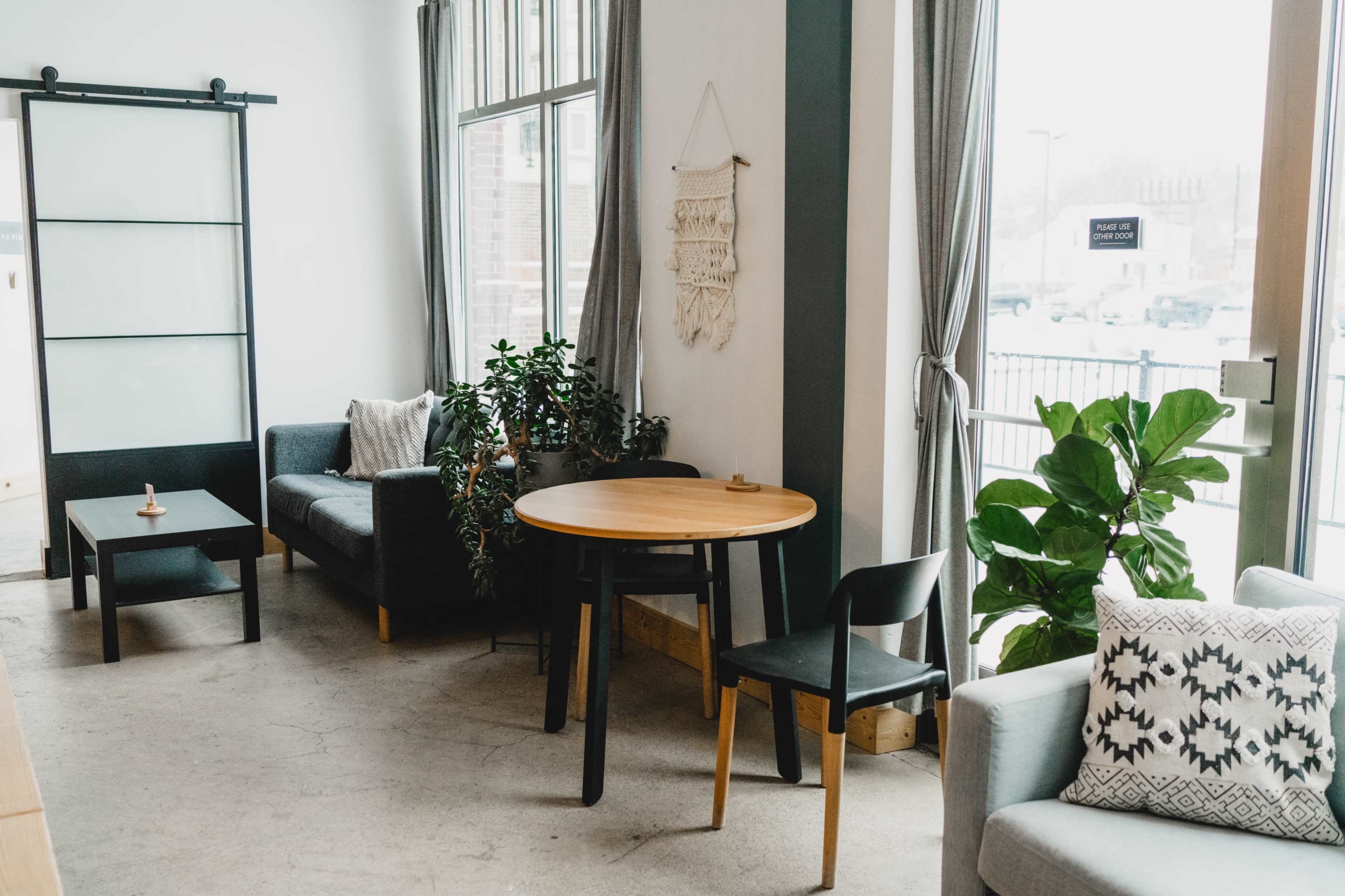 A contemporary indoor seating area with two couches, a round table, and plants against large windows.
