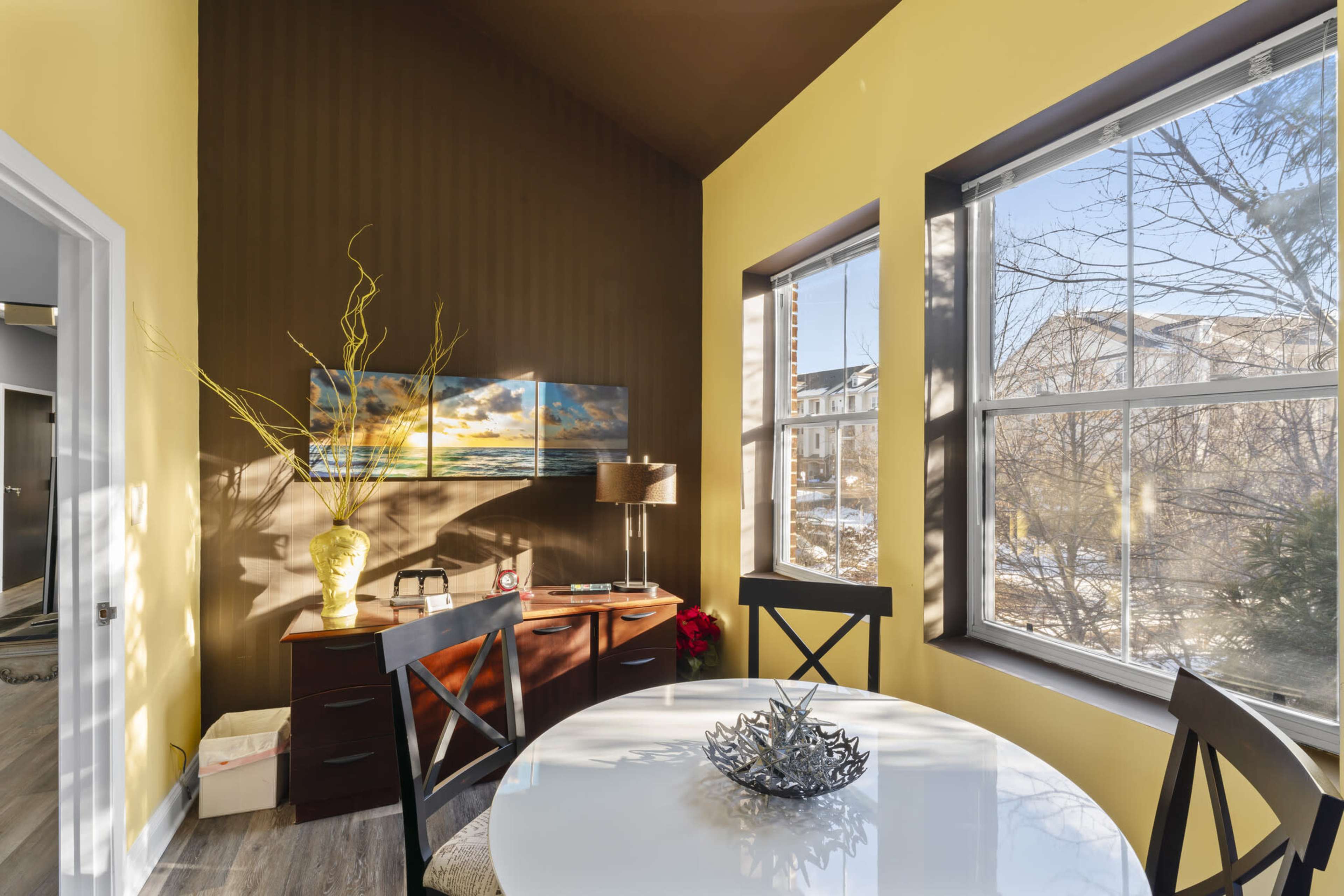 A small dining area features a round white table and dark wood chairs, with yellow and brown walls, a colorful painting, and windows allowing natural light to enter.