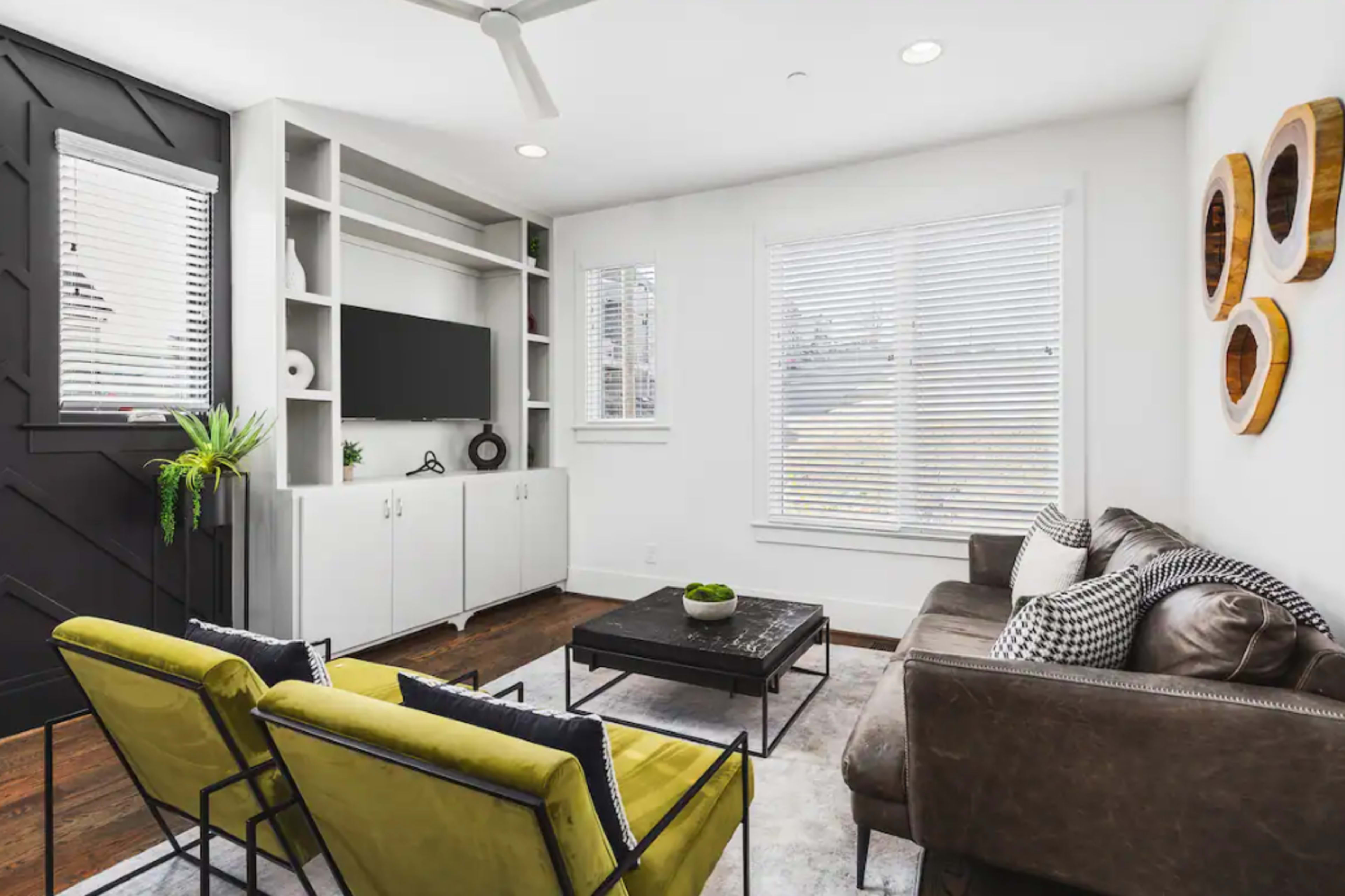 A modern living room featuring a dark accent wall, a television on a white shelving unit, a gray sofa, and two yellow chairs around a coffee table.