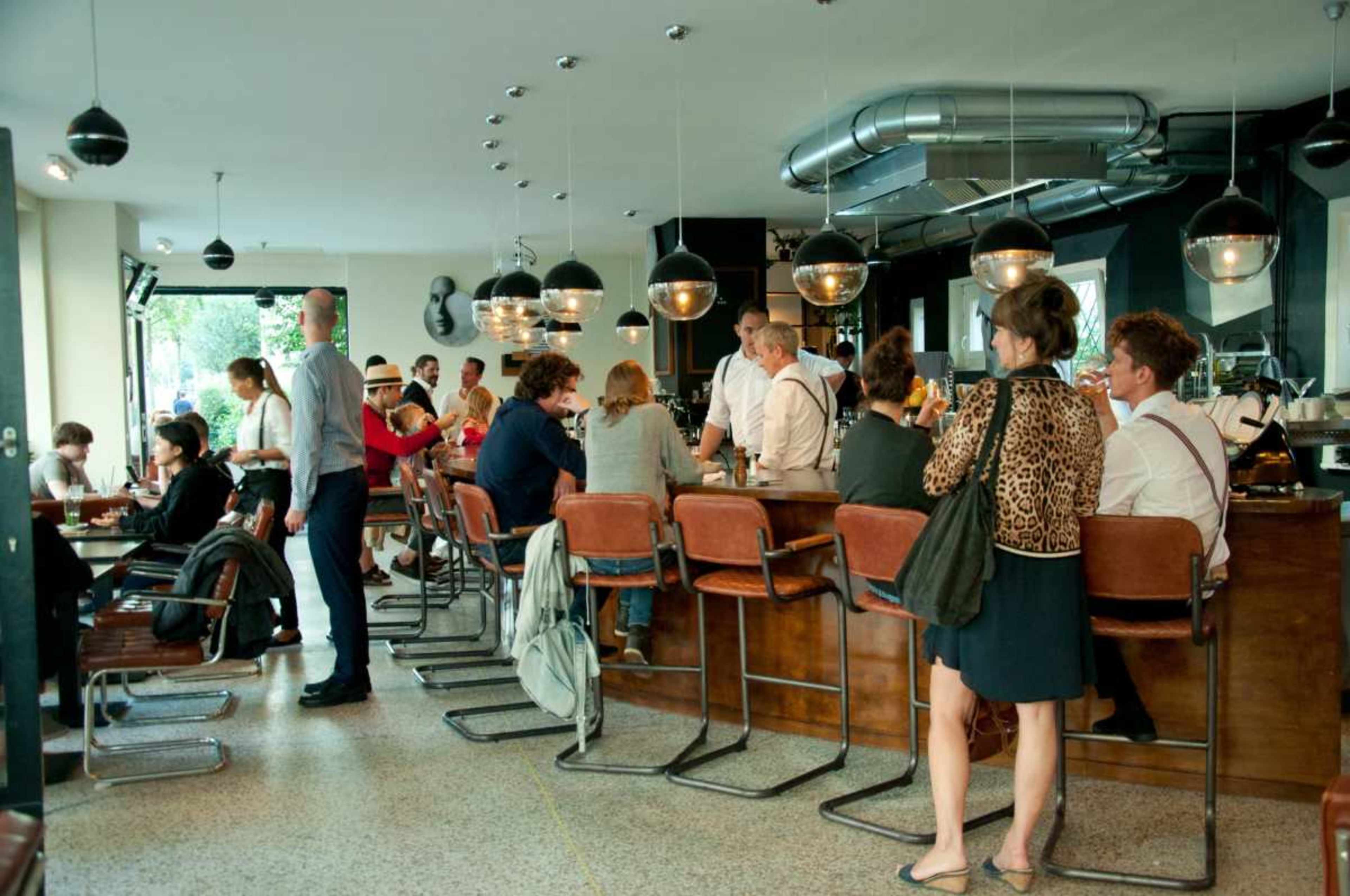 The image shows a bustling café interior with patrons seated at a long bar and others mingling, surrounded by modern decor and lighting fixtures.