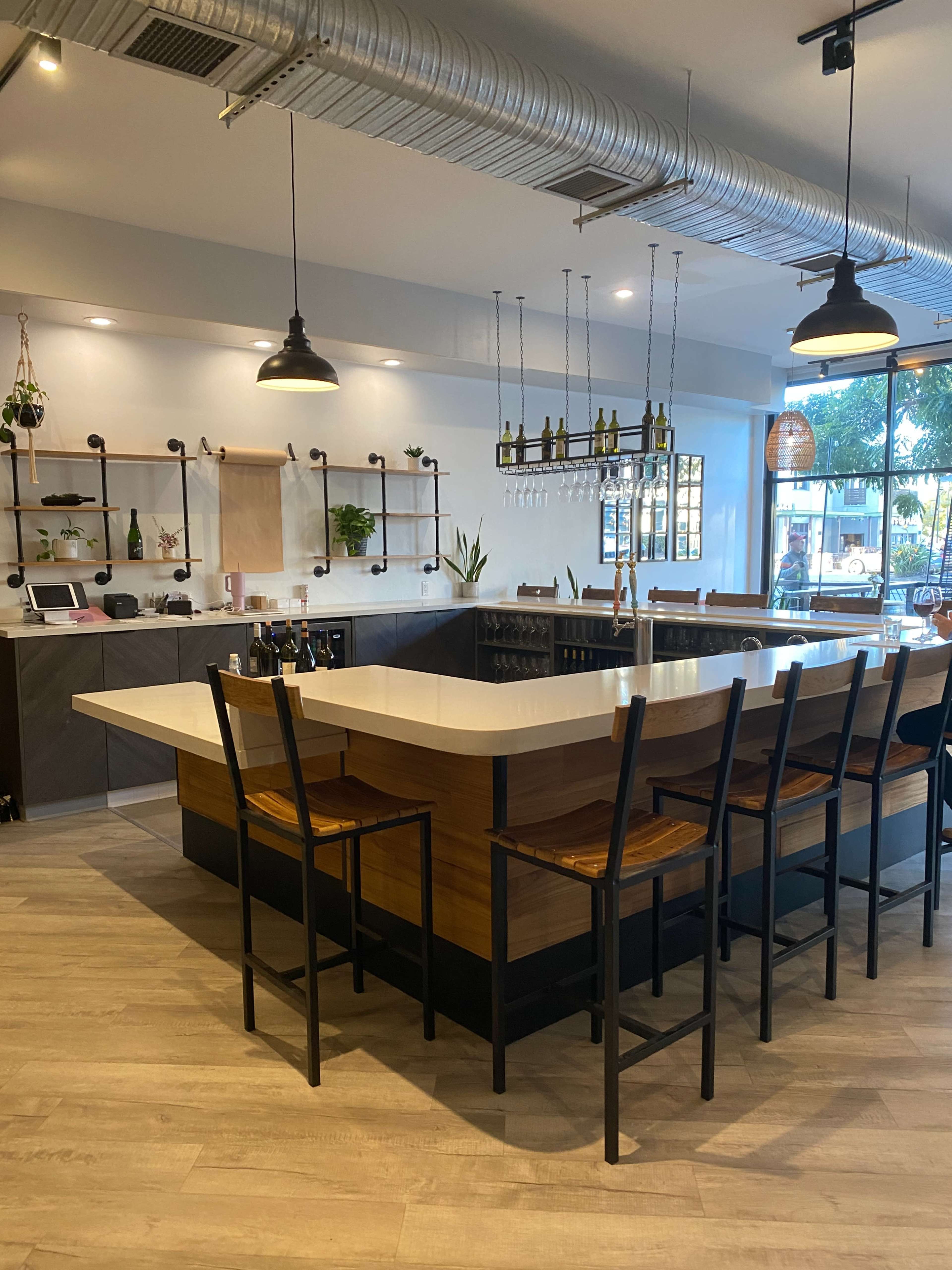 The image shows a modern restaurant interior featuring a large island bar with high stools, wooden accents, and shelving with plants and bottles.