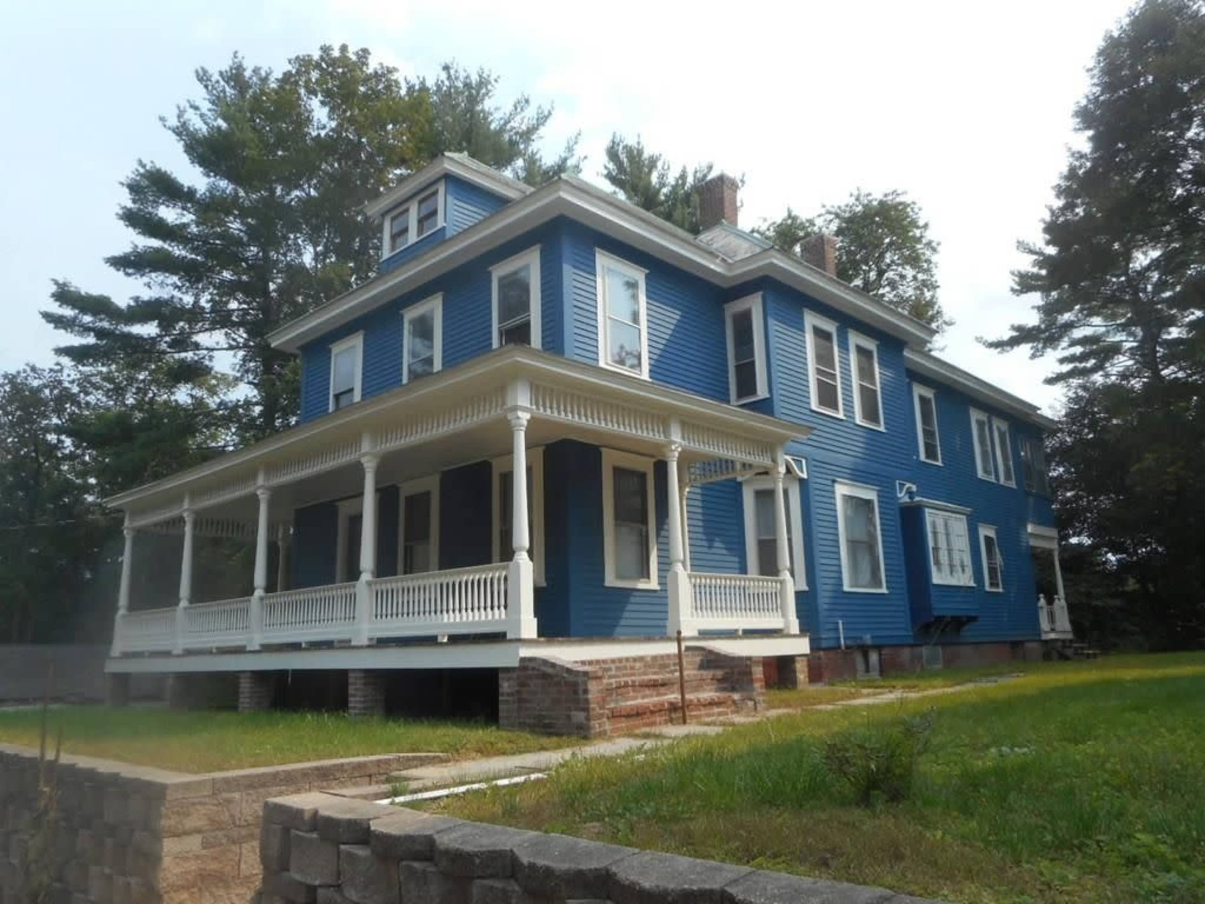 A large blue house with a wraparound porch and multiple gabled roofs, surrounded by trees and a grassy area.