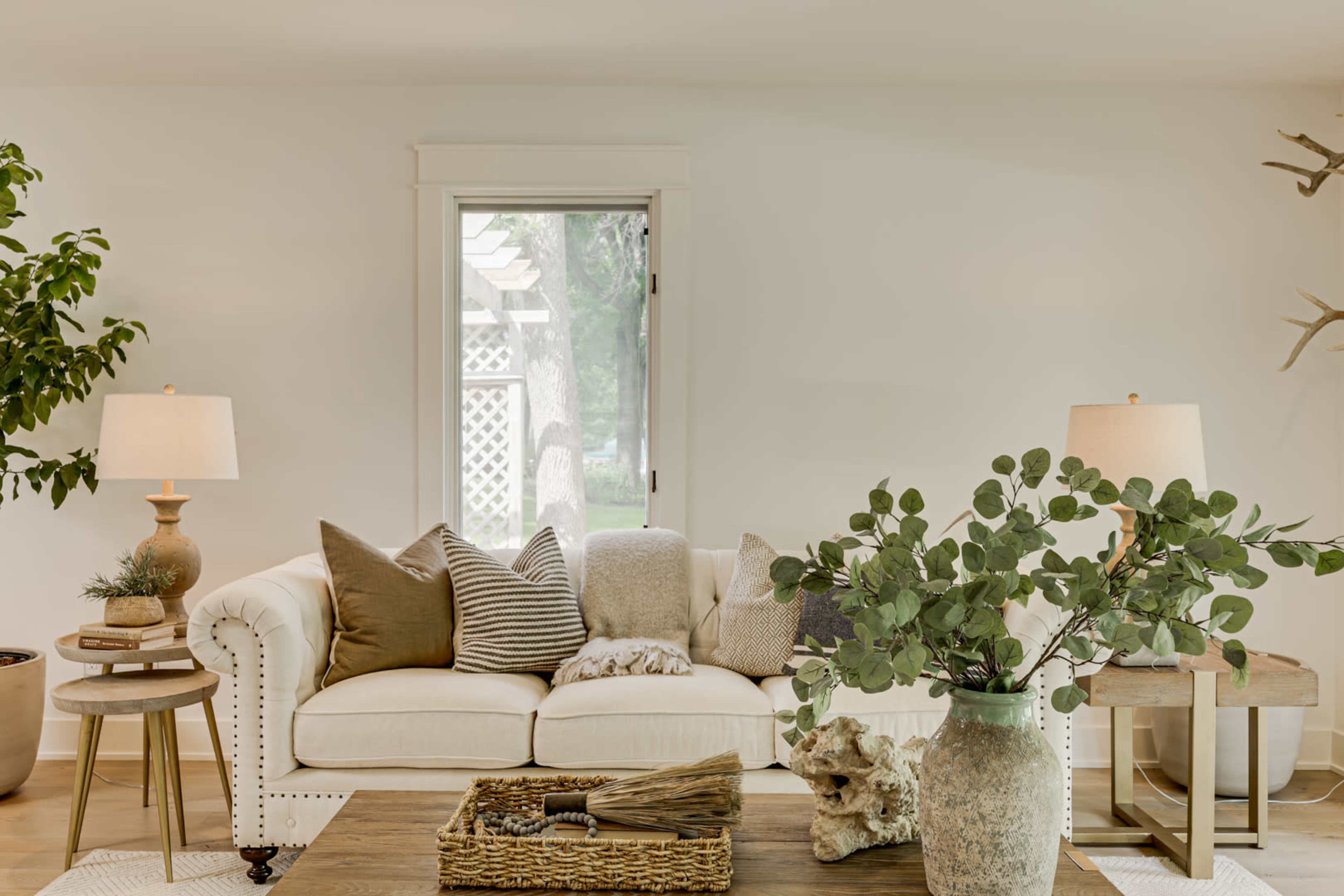 A cozy living room with a light-colored sofa adorned with various cushions, a wooden coffee table, and plants beside two lamps.