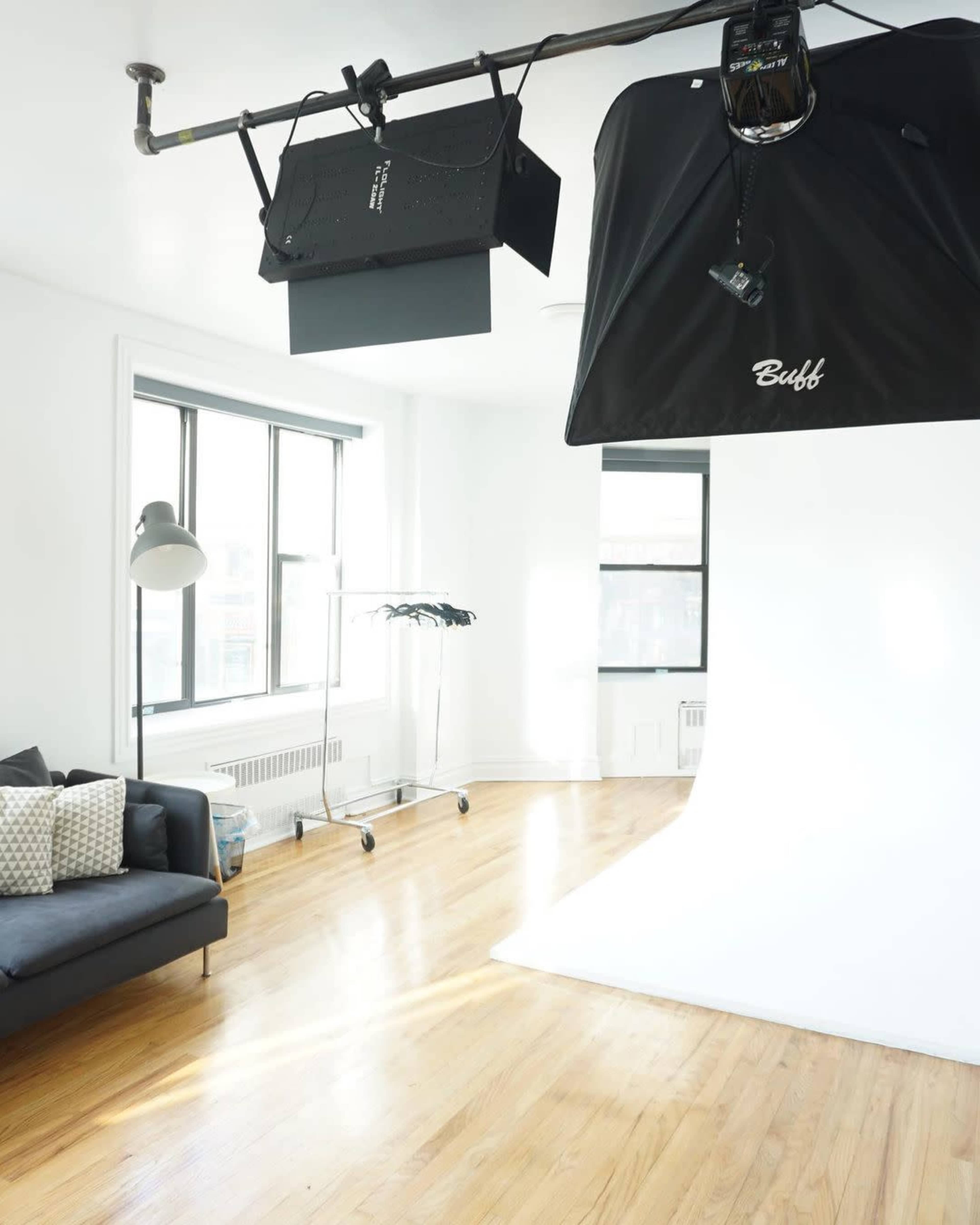 A well-lit photography studio with a large white backdrop, a light setup suspended from the ceiling, and a couch beside a clothing rack.