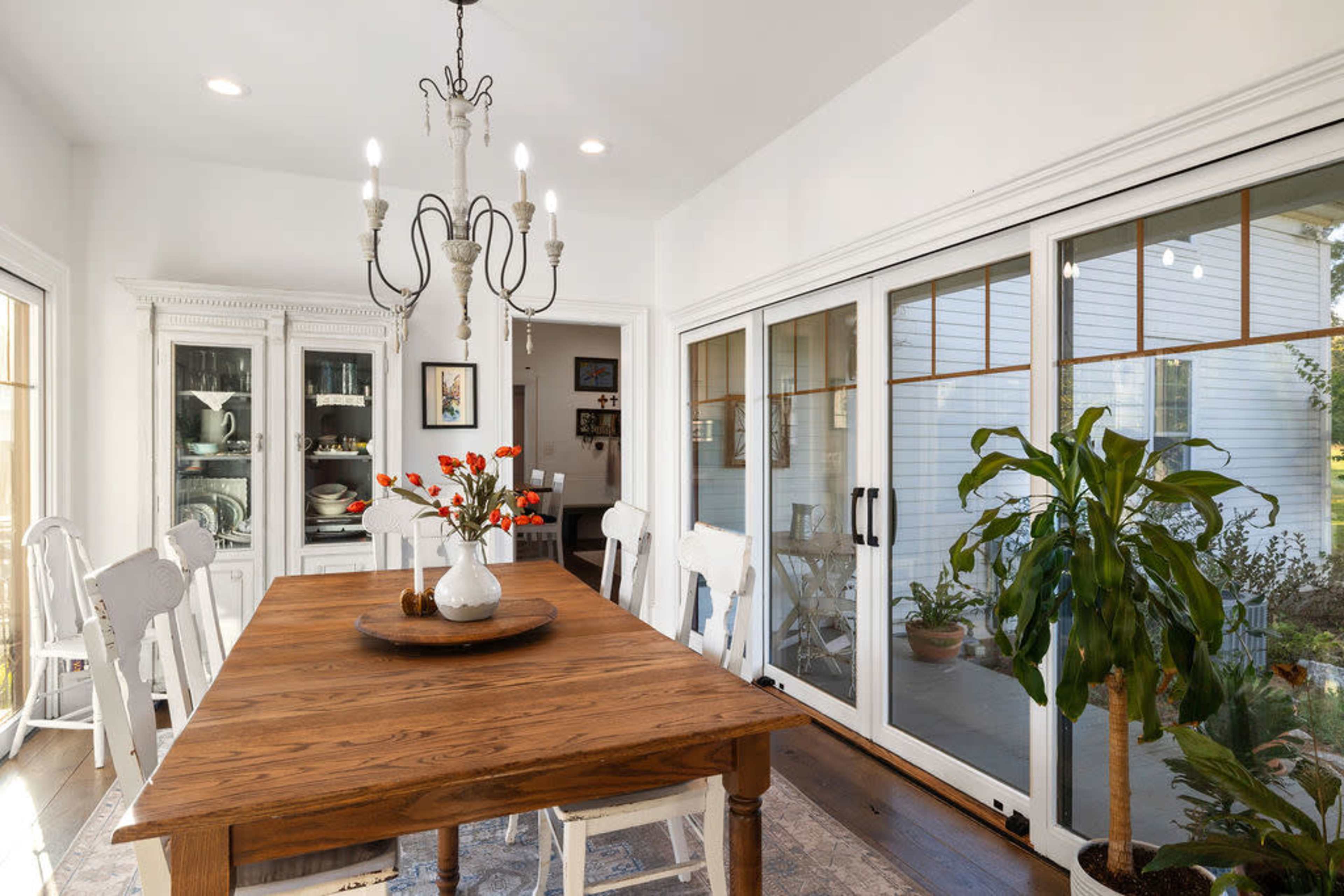 The image shows a bright dining room featuring a wooden table with a floral centerpiece, surrounded by white chairs, and large glass doors leading to an outdoor area.