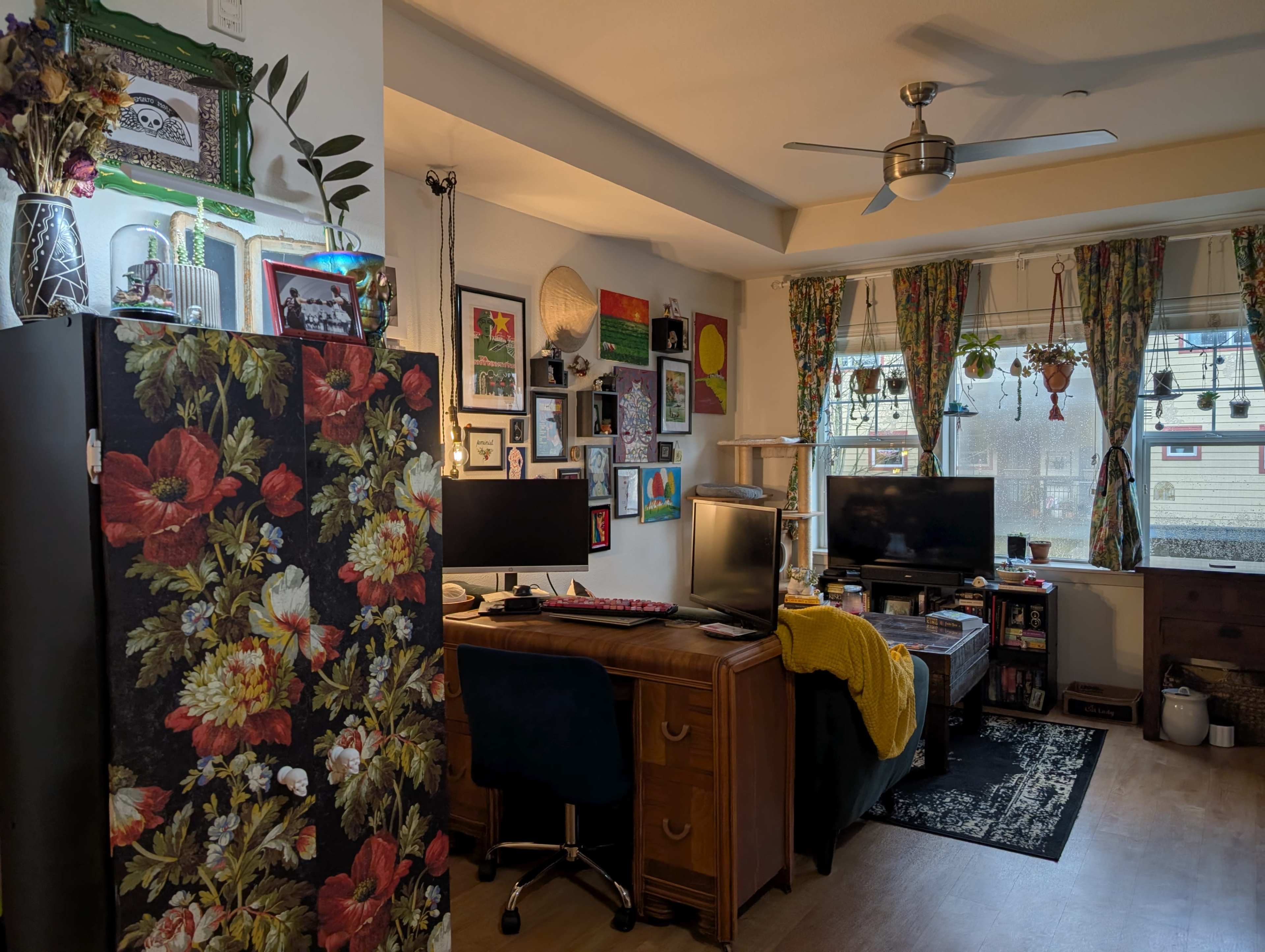 A cozy living room features a desk with dual monitors, a dark floral-patterned cabinet, and a window adorned with colorful curtains, surrounded by various decorations and wall art.