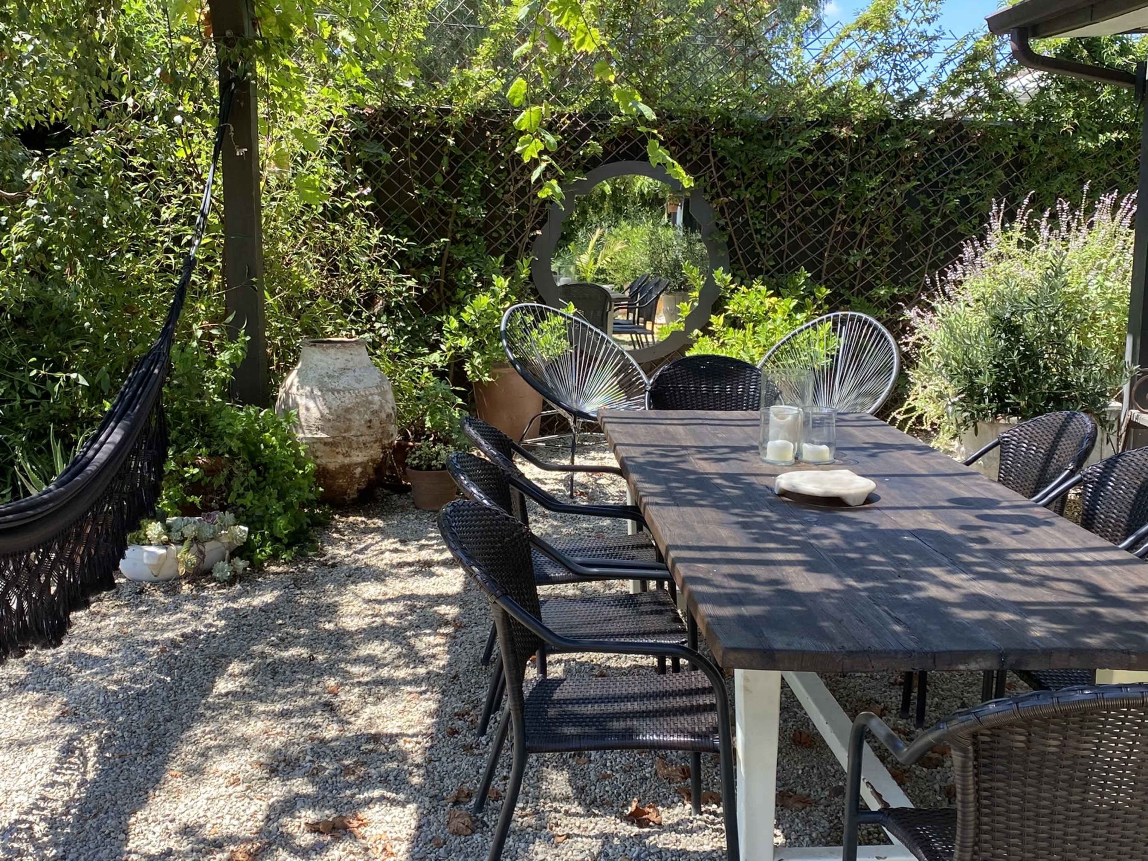 A wooden table surrounded by black chairs is set in a gravel courtyard, with green plants and a hammock visible in the background.