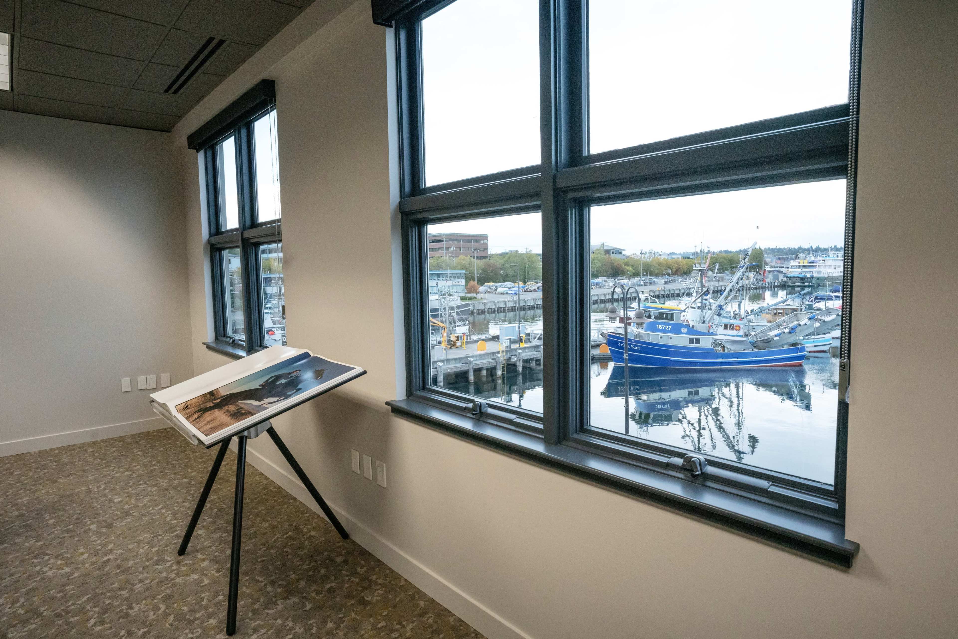The image shows a room with large windows overlooking a harbor filled with boats, while an easel displaying a photograph stands nearby.