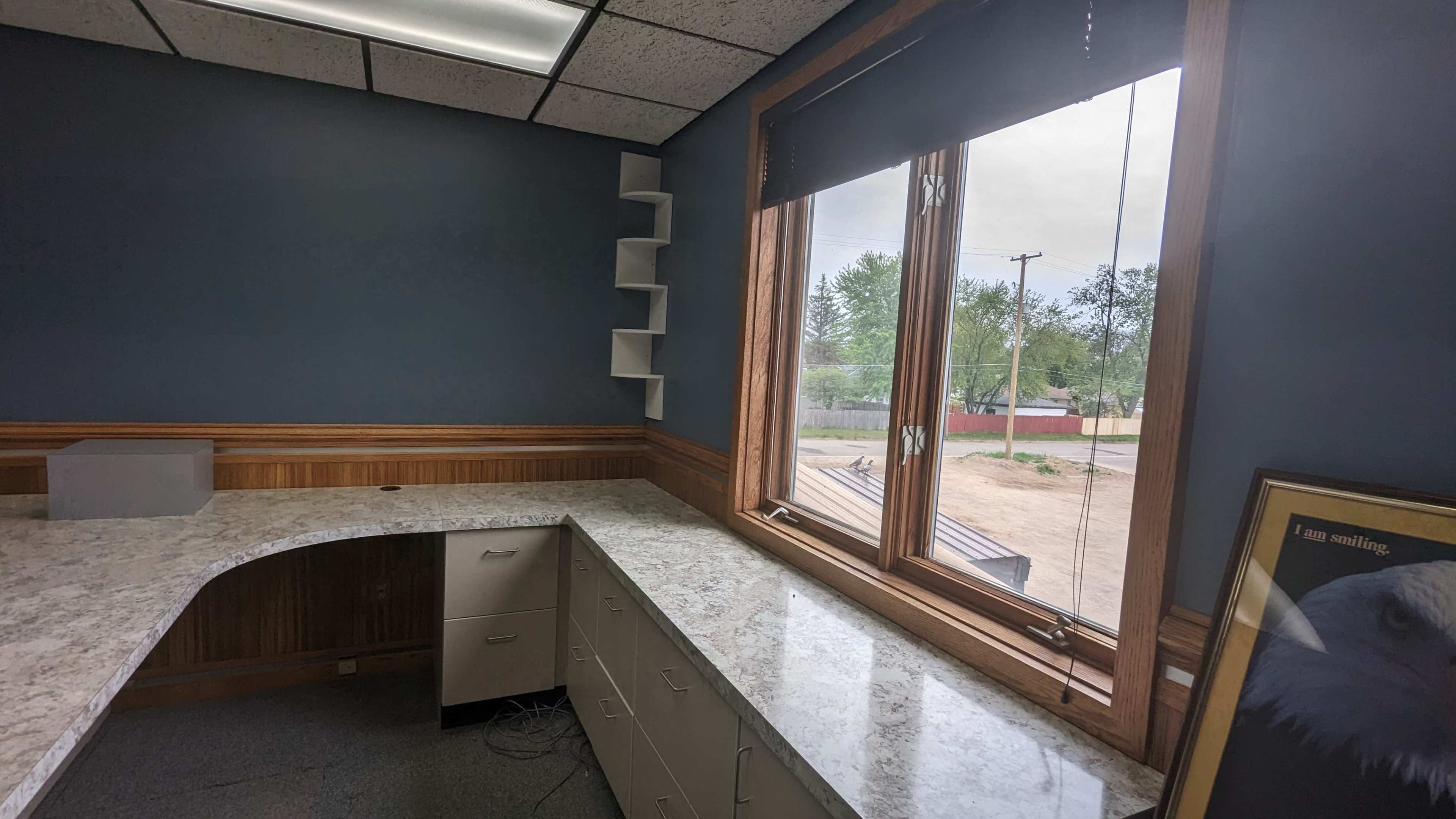 The image shows a corner of an office with a granite countertop, cabinets beneath, and a large window overlooking a rural landscape.