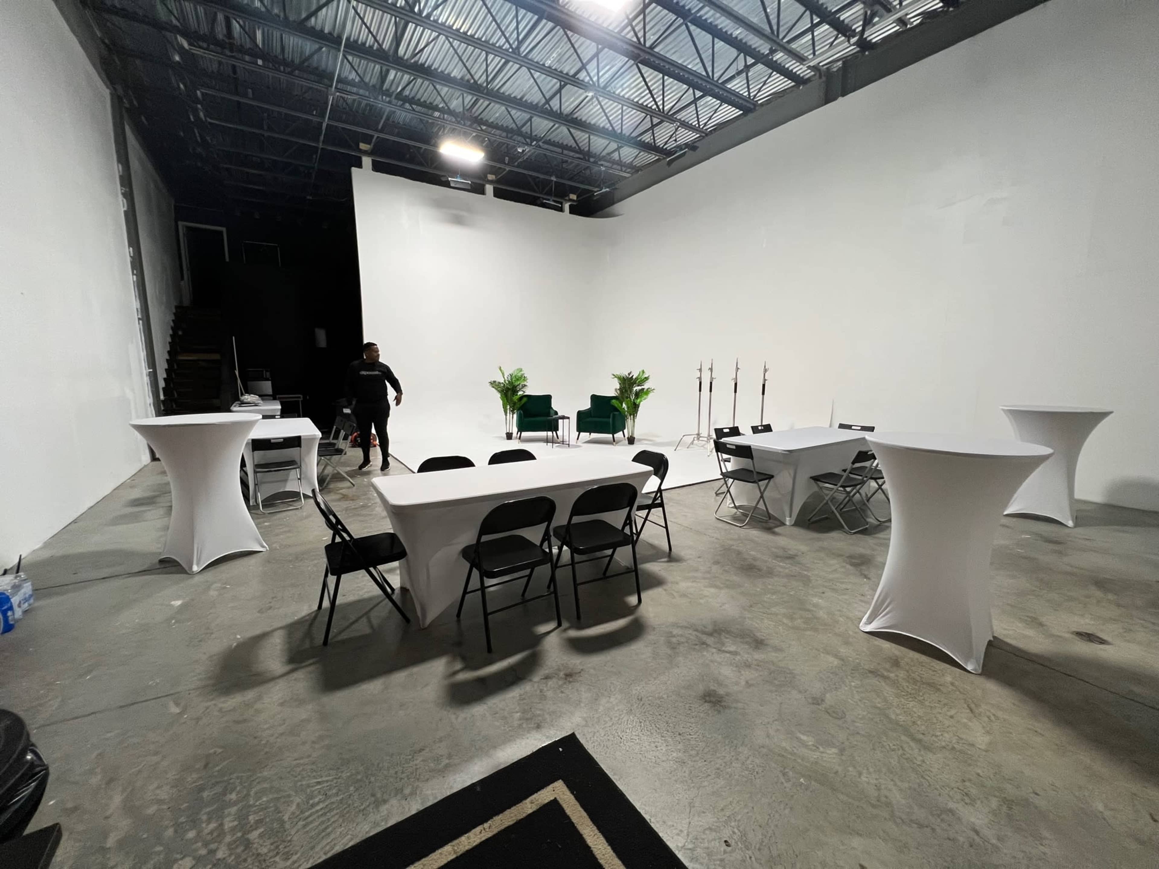 An empty event space with multiple white tables and chairs arranged, a plain white wall in the background, and a few green plants.