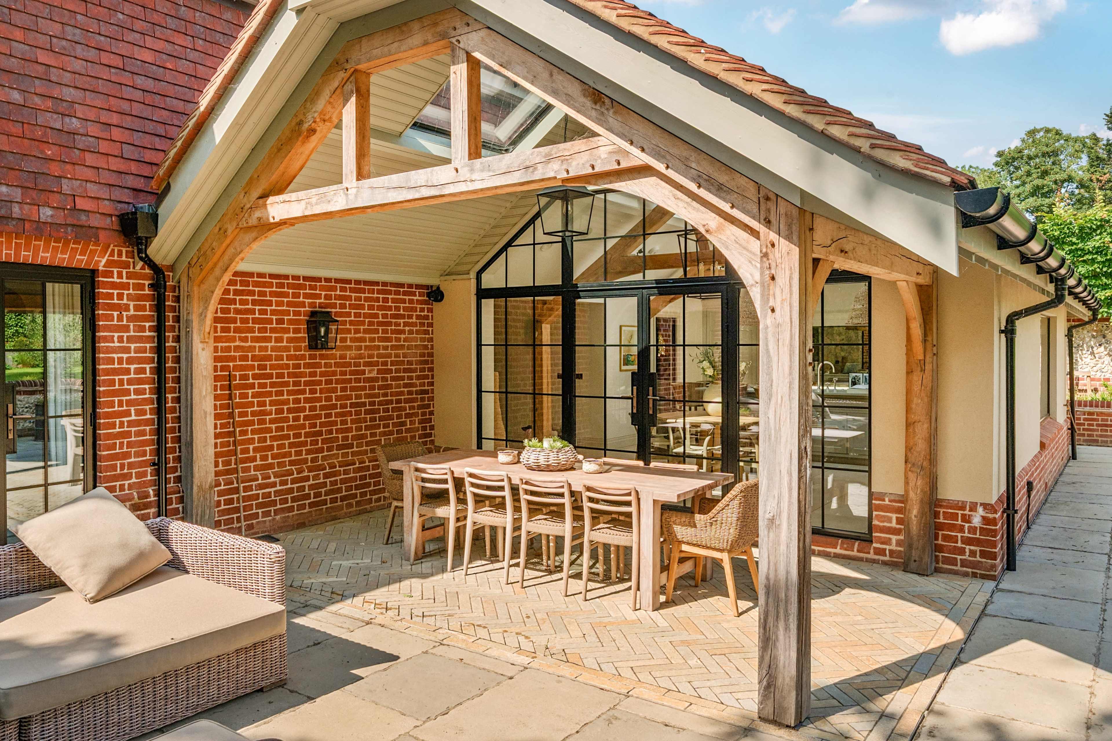 The image shows a patio area with a wooden frame, featuring a dining table surrounded by chairs and a comfortable sofa, adjacent to a brick wall building.