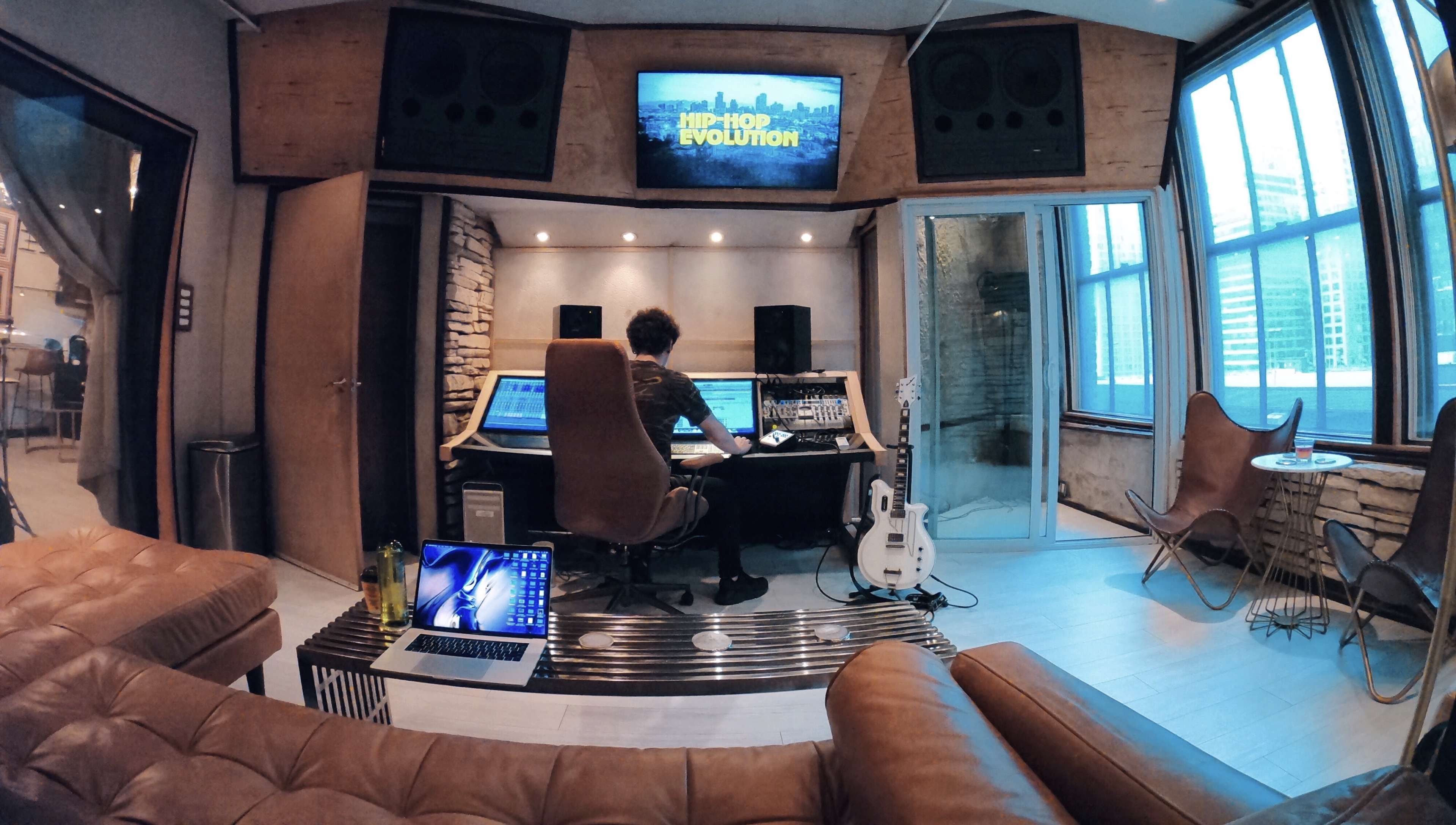 A person sits at a mixing console in a modern recording studio with large speakers and a television displaying "HIP-HOP EVOLUTION."