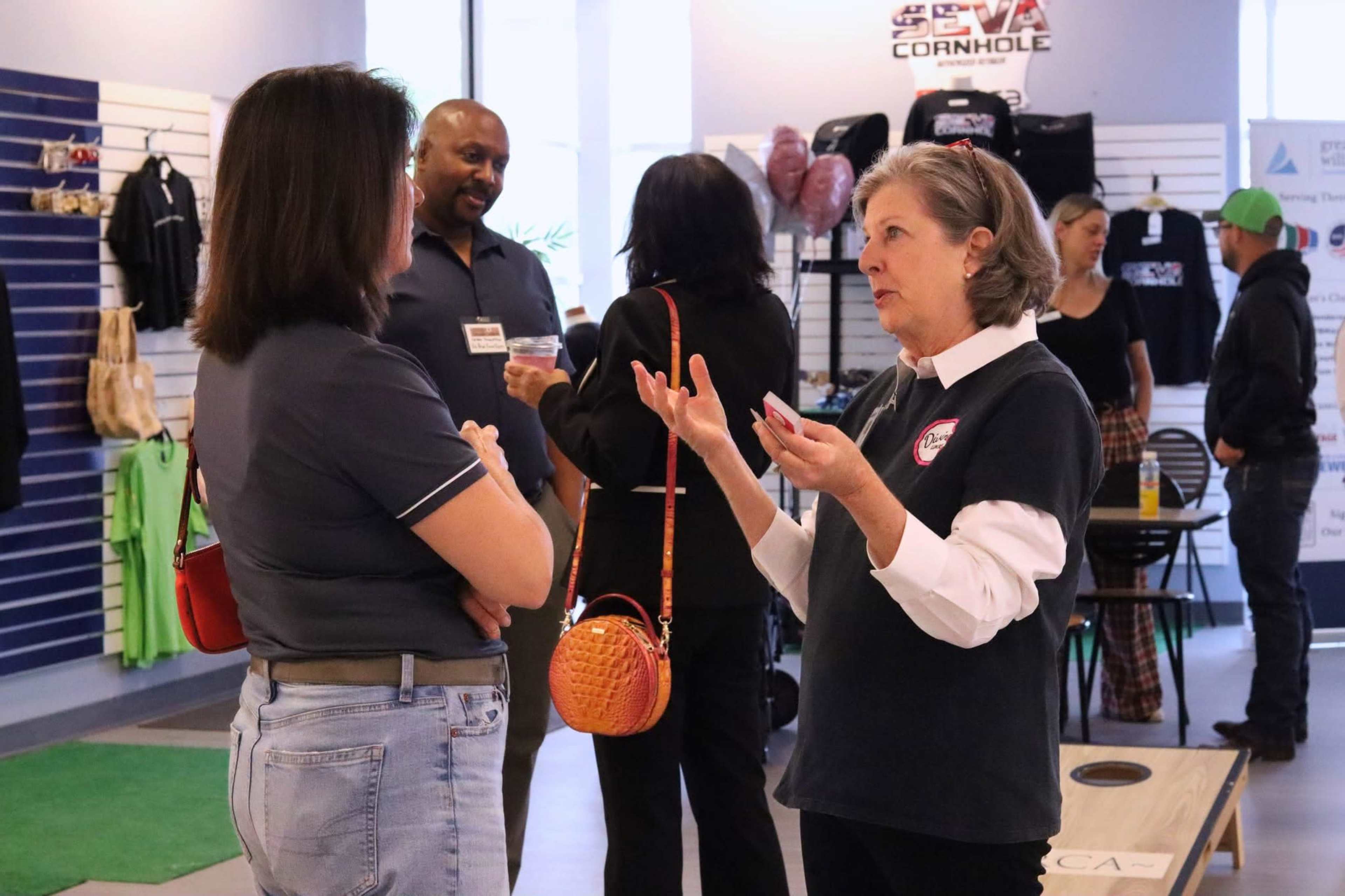 Two women are engaged in conversation in a shop filled with various merchandise, while other individuals socialize in the background.