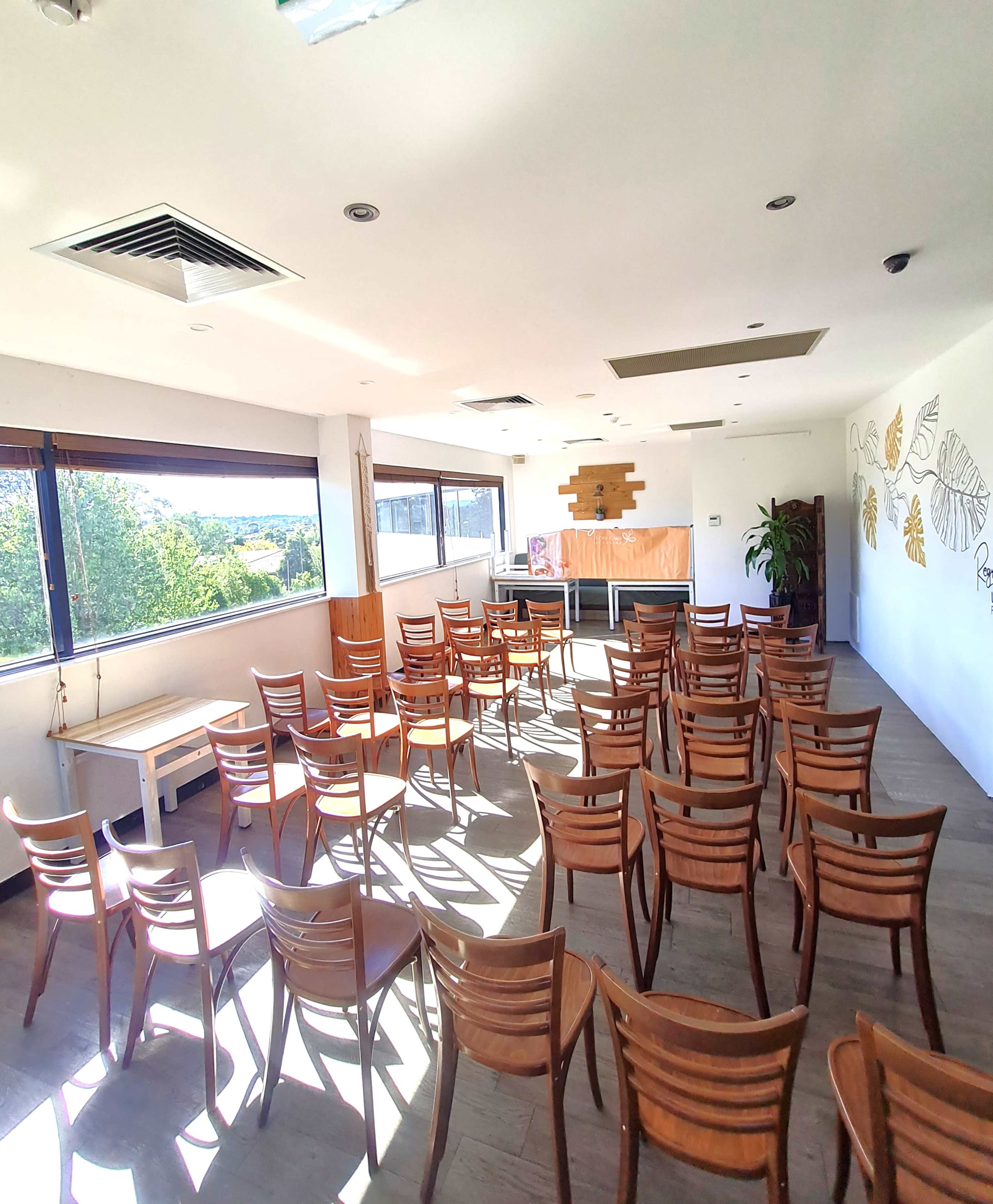 The image shows a well-lit room arranged with wooden chairs facing a presentation area, featuring large windows with views of greenery outside.