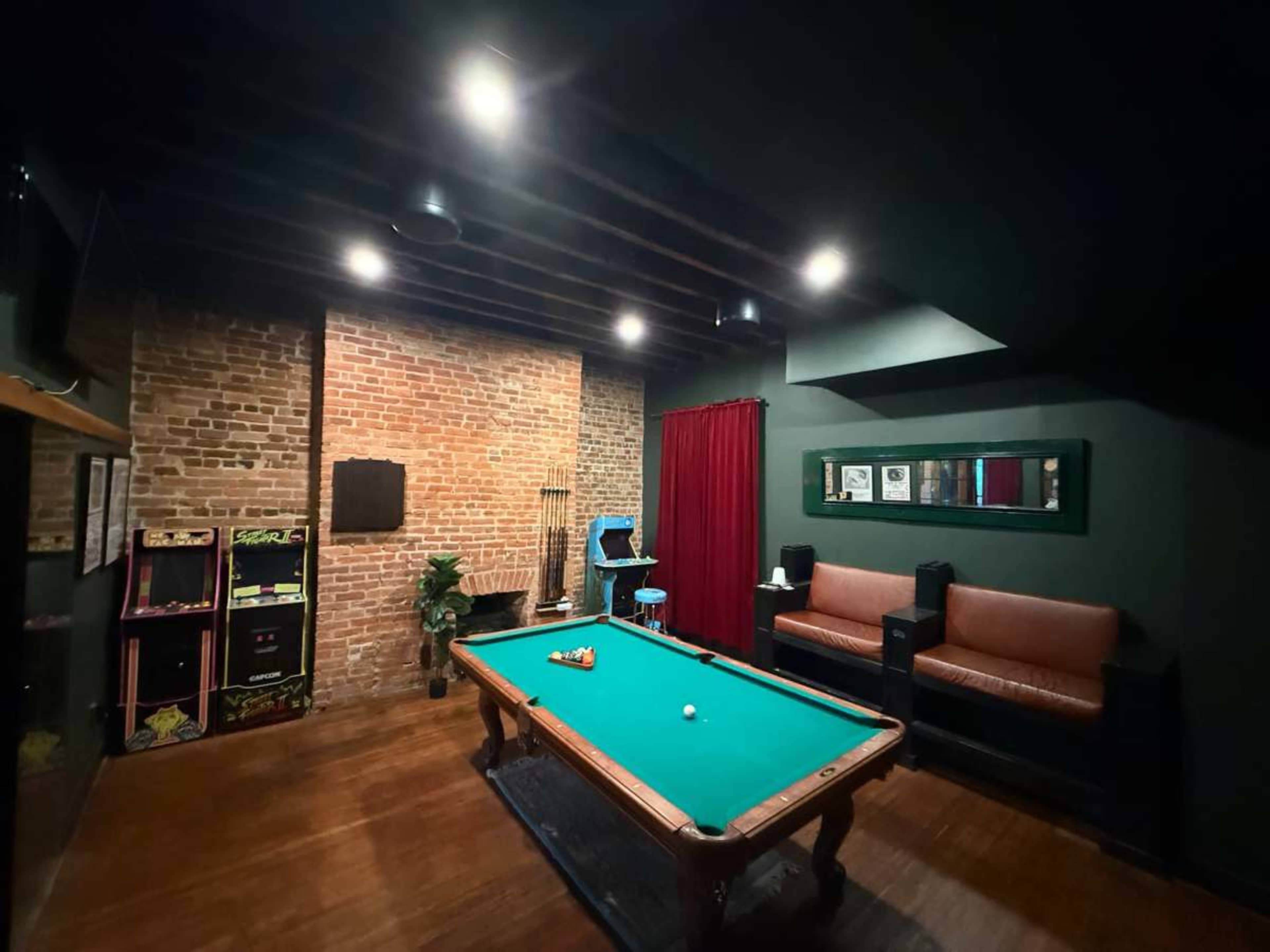 The image shows a game room featuring a pool table, a brick wall, a couple of leather couches, and vintage arcade machines.
