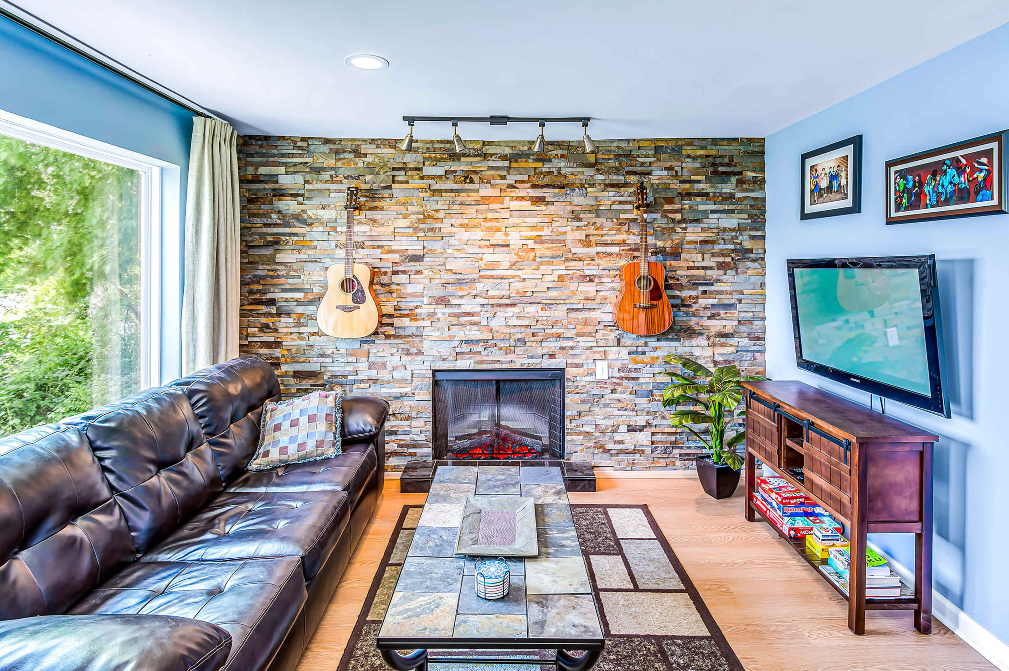 A living room with a stone feature wall, two acoustic guitars hanging on the wall, a black leather couch, a modern television, and a decorative coffee table.