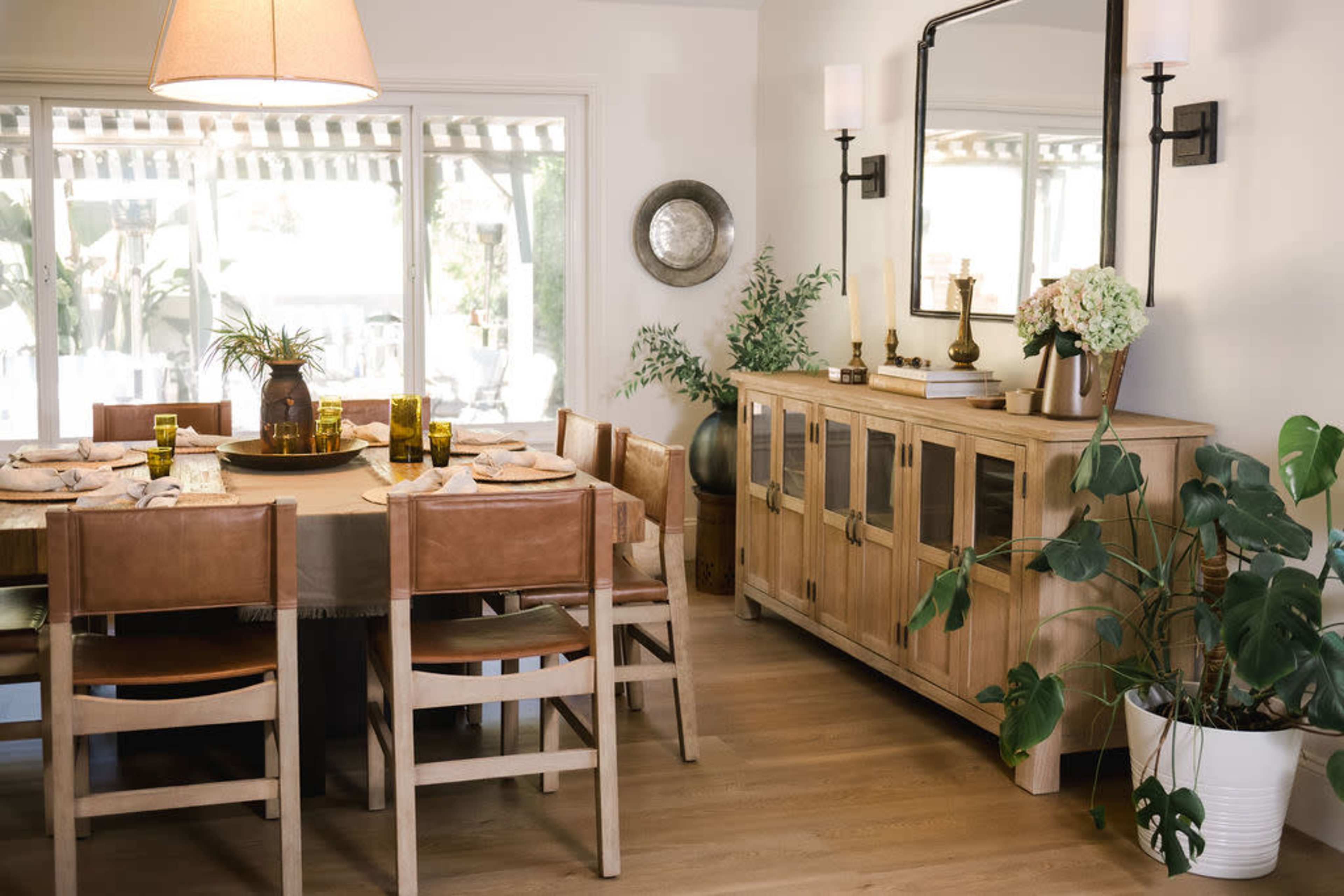 A dining room features a large wooden table surrounded by leather chairs, a sideboard with glass doors, and plants enhancing the decor.