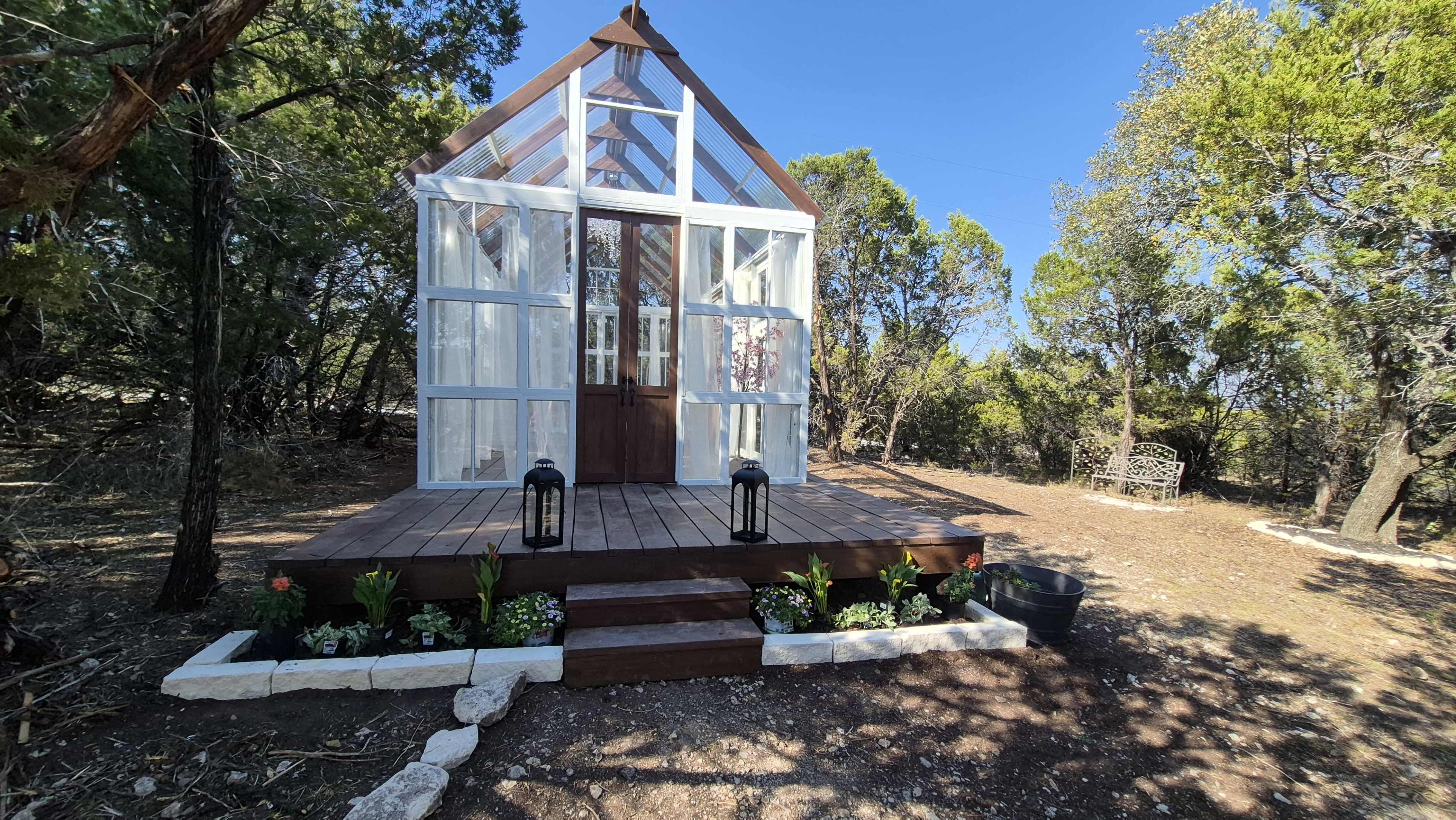 A glass-fronted greenhouse with wooden accents is situated among greenery, featuring a wooden deck, lanterns, and landscaped flower beds.