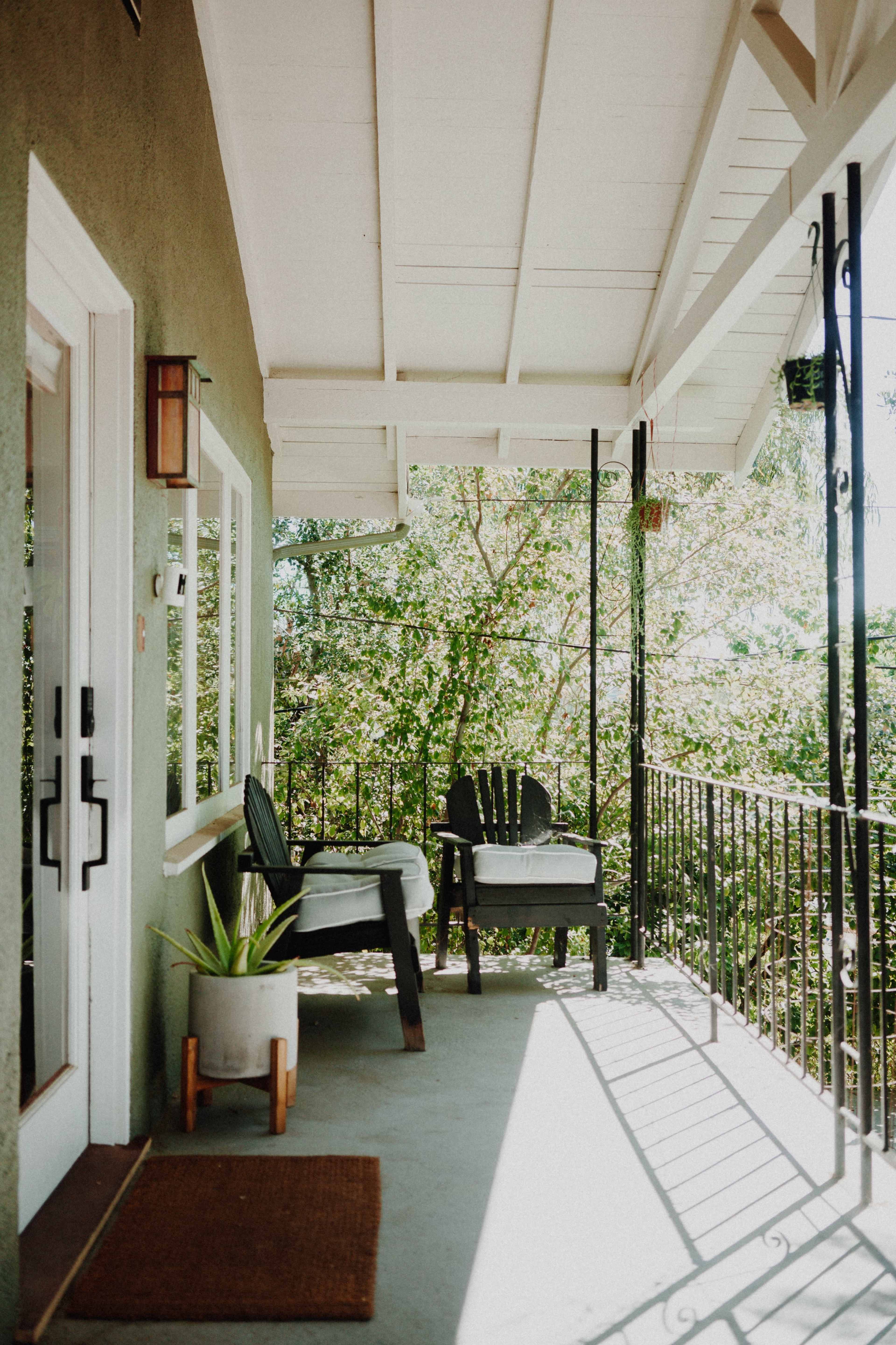 A covered porch features two black chairs with white cushions, a small potted plant, and a railing overlooking a lush green landscape.