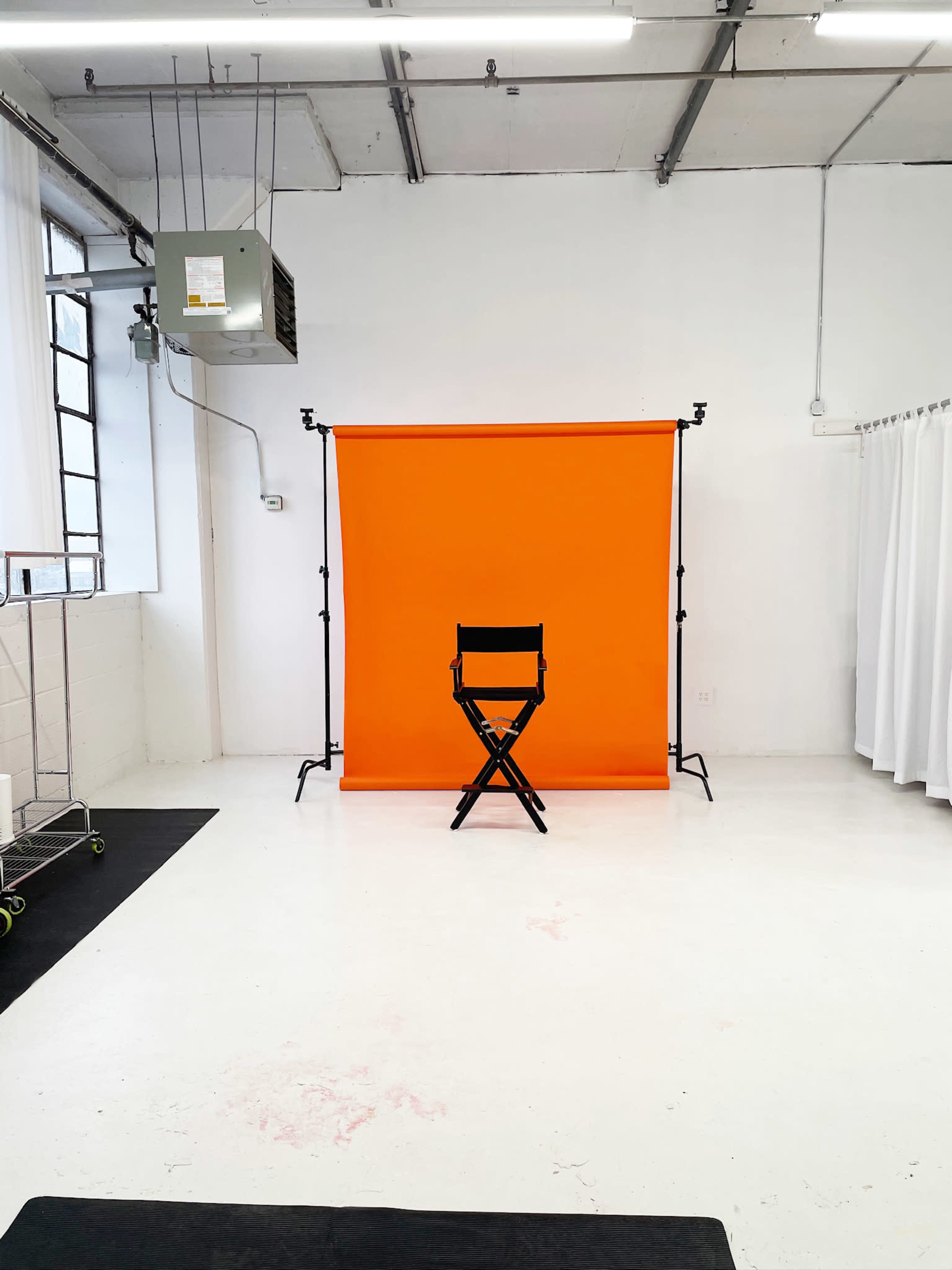 The image shows a photography studio with a black director's chair positioned in front of an orange backdrop, set against a bright white floor and walls.
