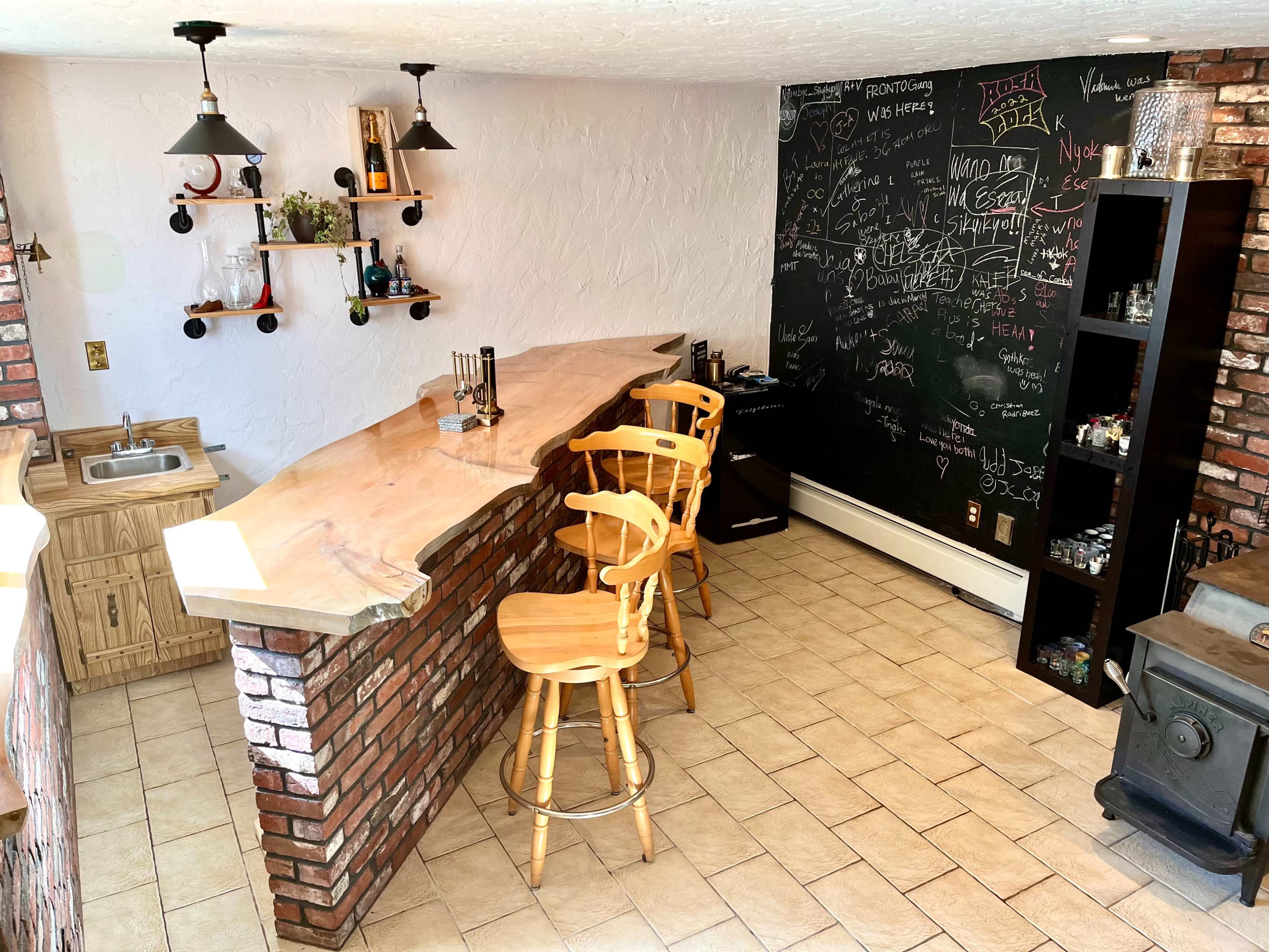 The image shows a home bar area featuring a wooden counter with three high stools, a sink to the left, a black chalkboard wall covered in writing, and shelves displaying various bottles.