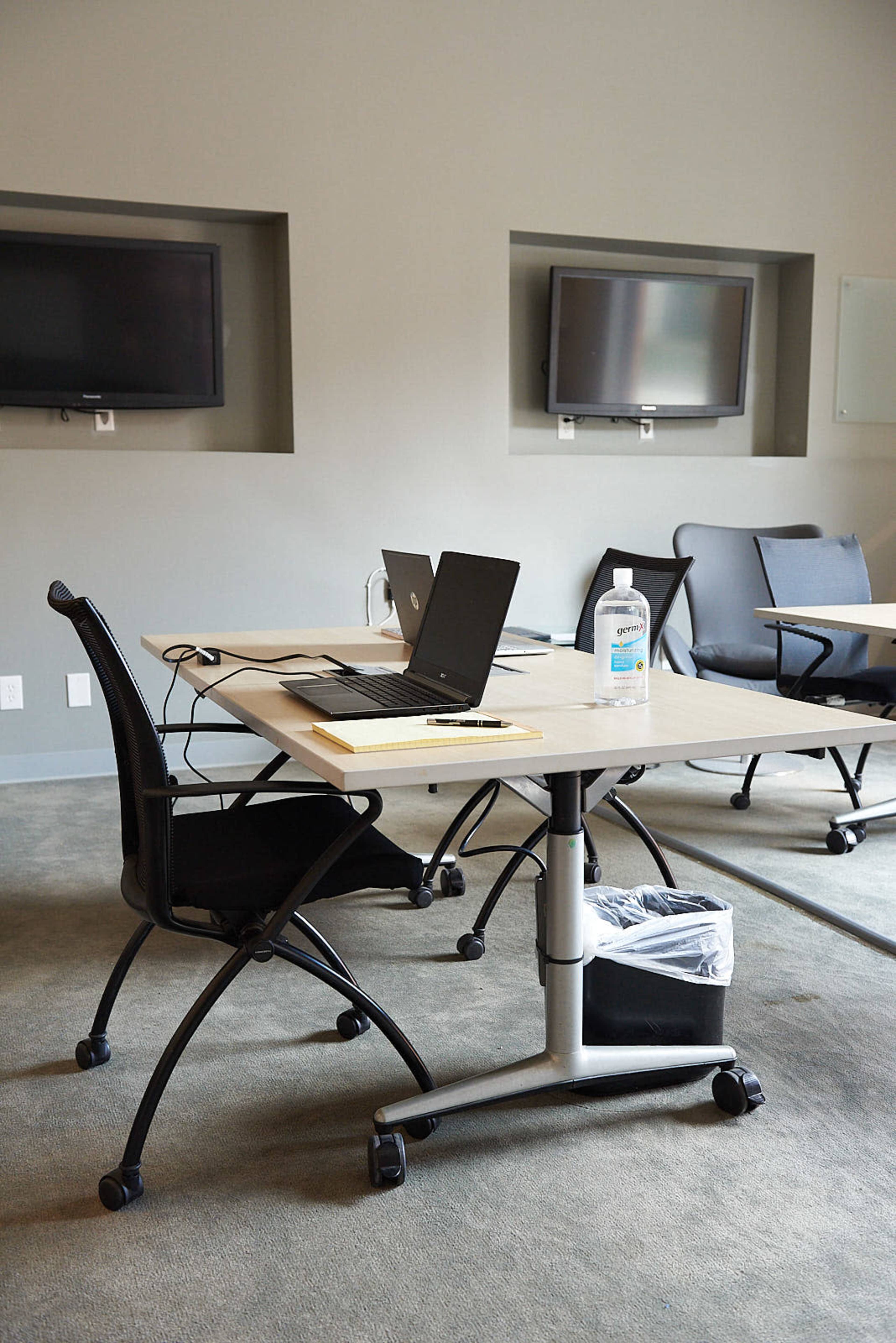 A conference room with two monitors mounted on the wall, a wooden table with laptops and a bottle of hand sanitizer, and a trash bin beneath the table.